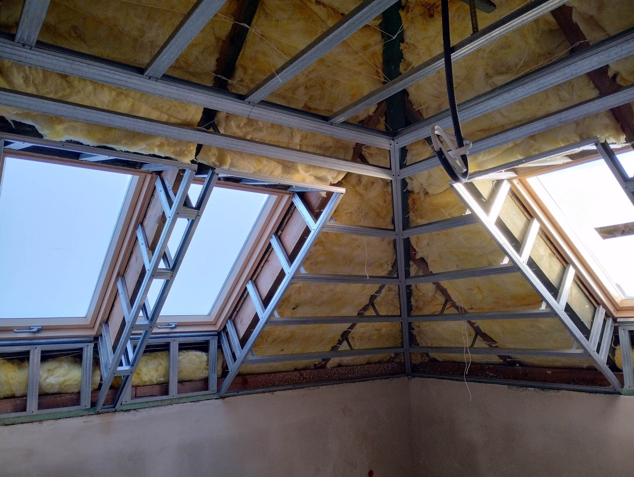 Montaż okien dachowych w konstrukcji szkieletowej z izolacją wełną mineralną. Widoczne metalowe profile i drewniane ramy okienne.