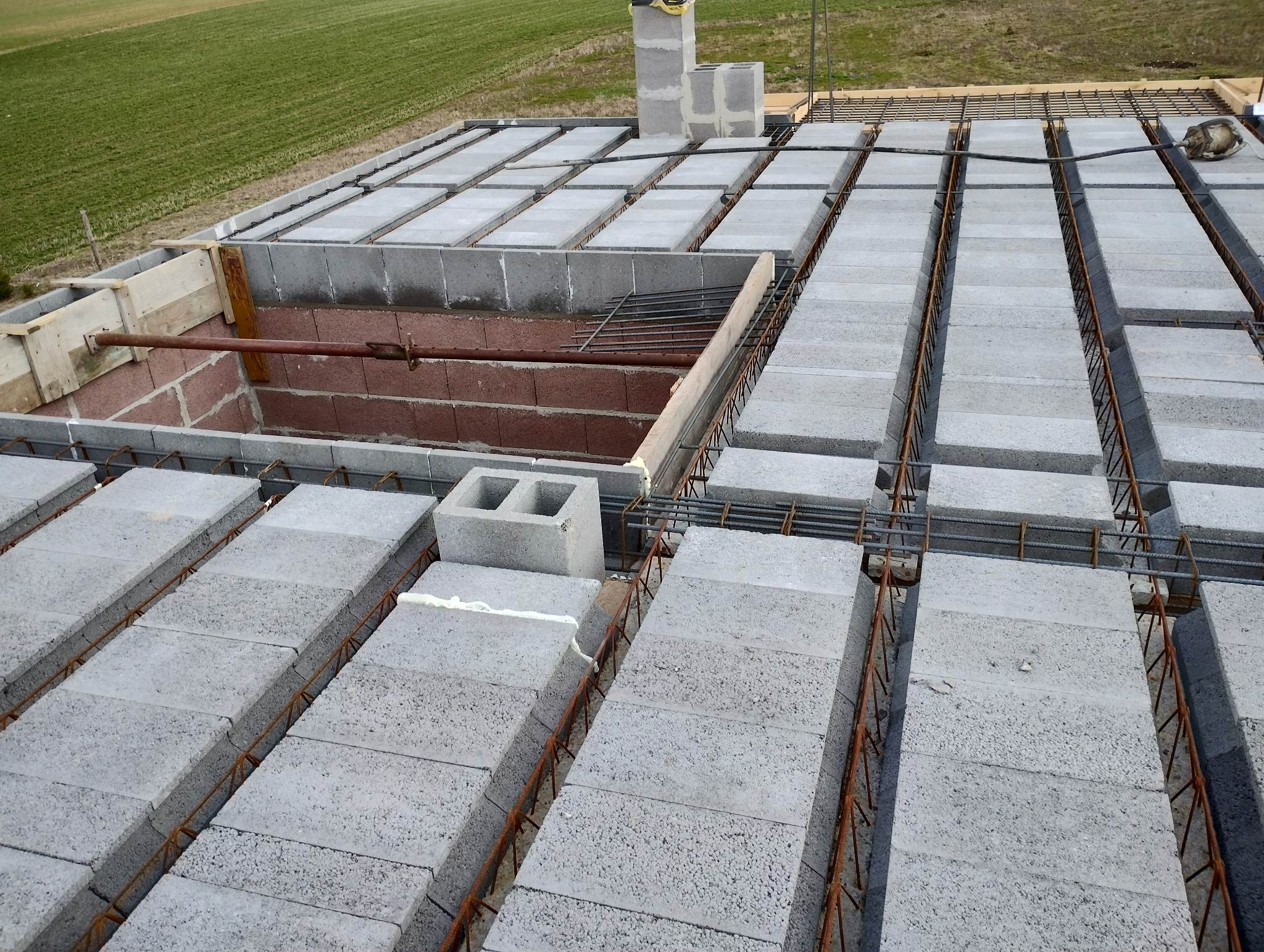 Wieniec stropowy z szalunkiem traconym, zbrojony stalą, wypełniony bloczkami betonowymi. Widoczne podparcie stemplami. Tło: zielone pole. Kąt ujęcia: z góry.