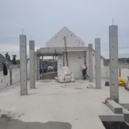 Budowa domu w trakcie murowania ścian z betonu komórkowego. Widoczne zbrojone słupy, ściany wznoszone z bloczków, rusztowania i pracownik budowlany.