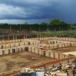 SOB-BUD - Drewniany szalunek fundamentów domu jednorodzinnego, przygotowany do zalania betonem. W tle ciemne chmury burzowe i rząd tuj.