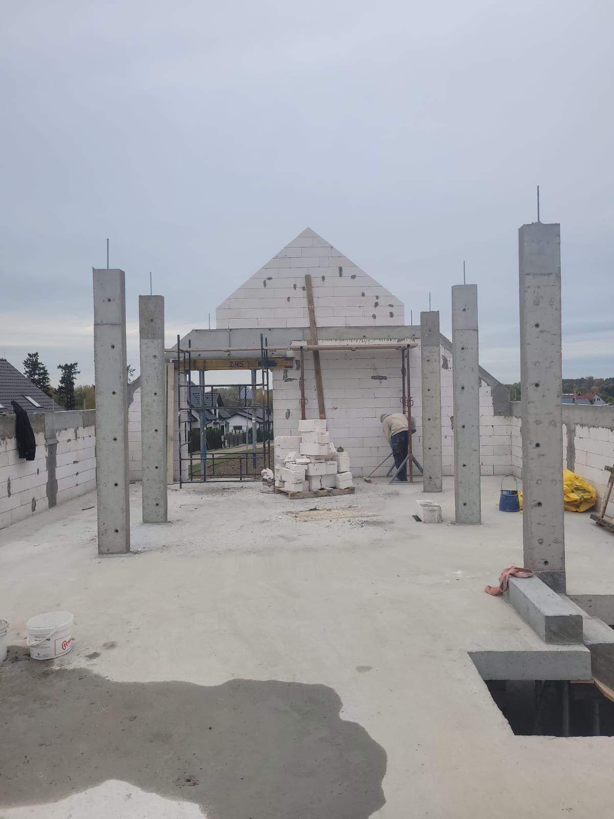 Budowa domu w trakcie murowania ścian z betonu komórkowego. Widoczne zbrojone słupy, ściany wznoszone z bloczków, rusztowania i pracownik budowlany.