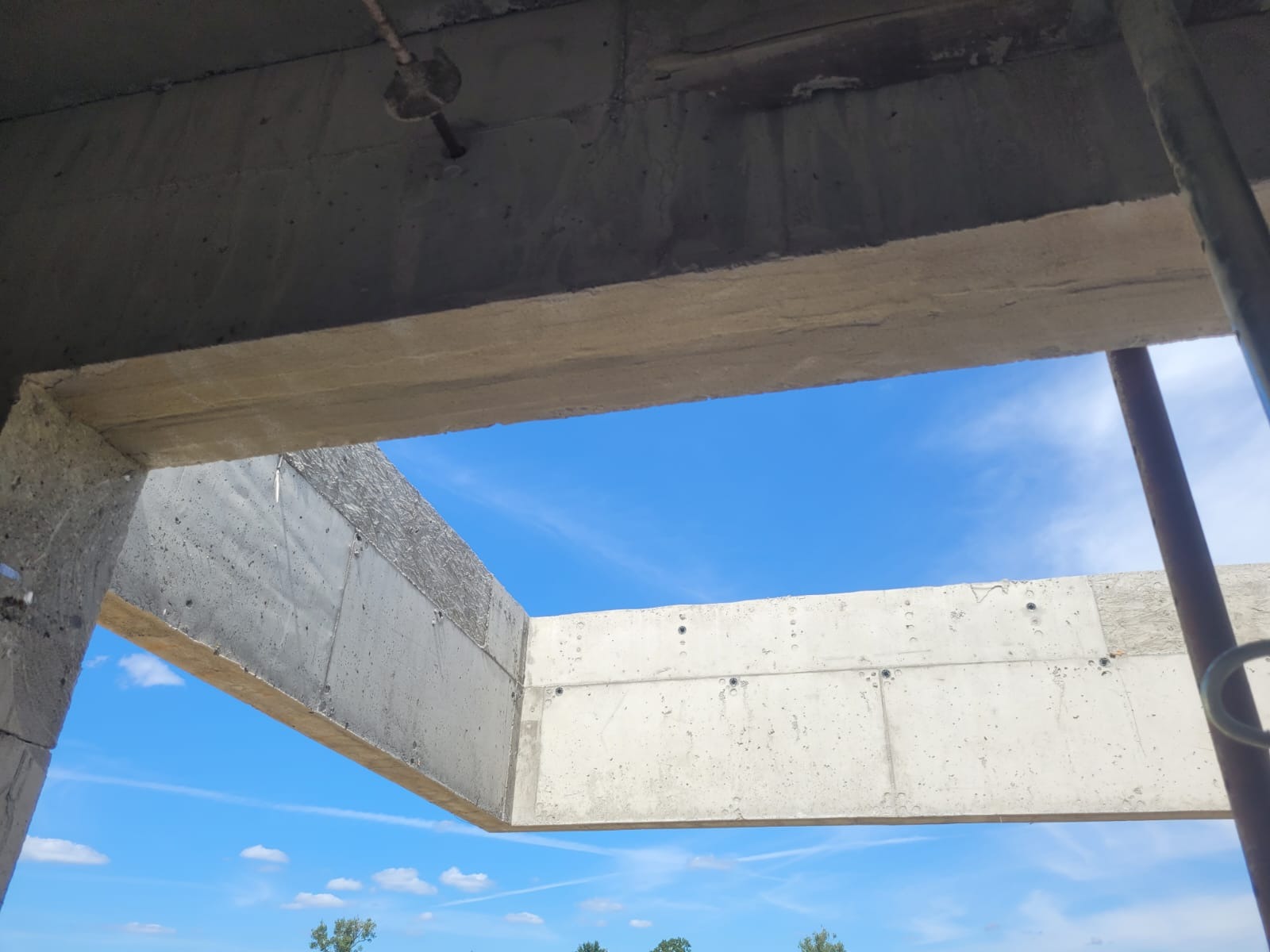 Surowa, betonowa konstrukcja budynku w trakcie budowy, widziana z dołu na tle błękitnego nieba. Widoczne ślady po szalunkach i elementy konstrukcyjne.