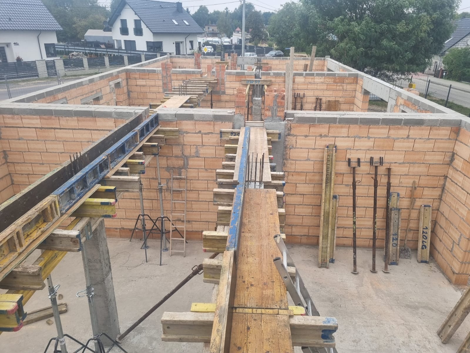 Budowa domu jednorodzinnego: murowane ściany z pustaków, szalunki pod wieniec, podpory i stemple. Widok z góry na plac budowy w słoneczny dzień.