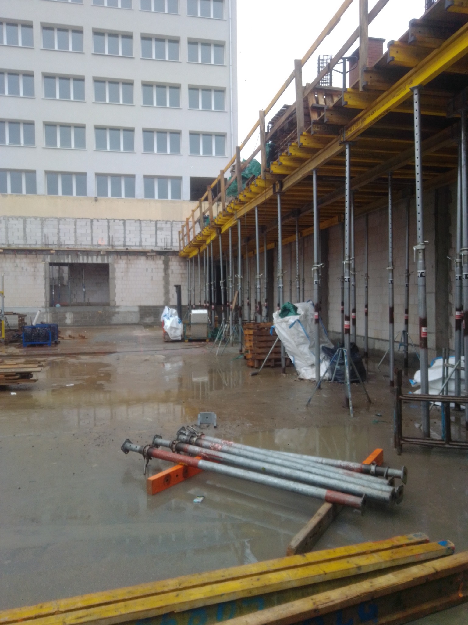 Budowa w toku: szalunki stropowe na podporach, widoczne zbrojenia i fragmenty murowanych ścian. Teren budowy mokry, po deszczu. W tle budynek wielopiętrowy.