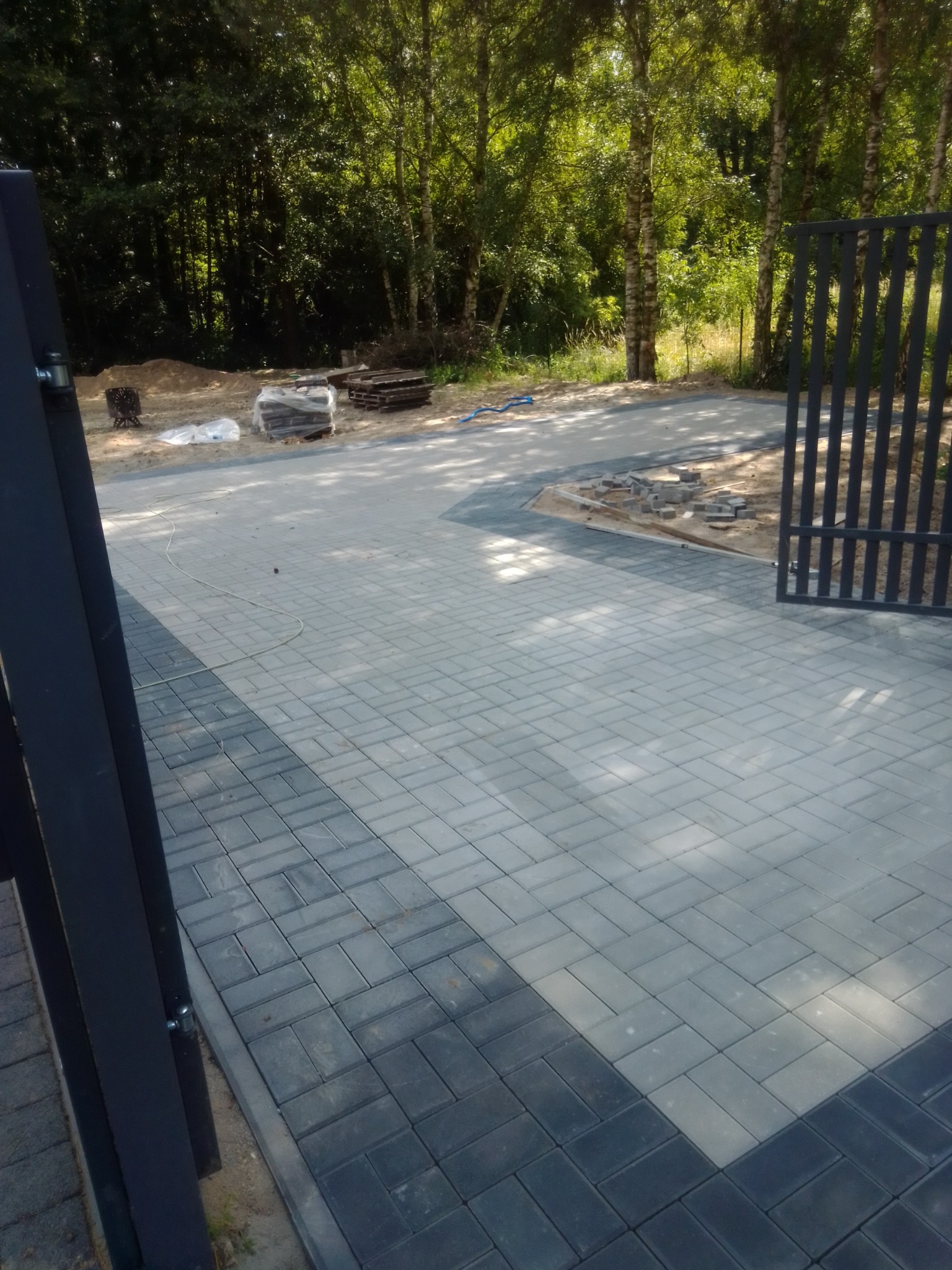 Ułożona kostka brukowa w dwóch odcieniach szarości tworząca wzór na podjeździe. W tle widoczne drzewa i elementy ogrodzenia.