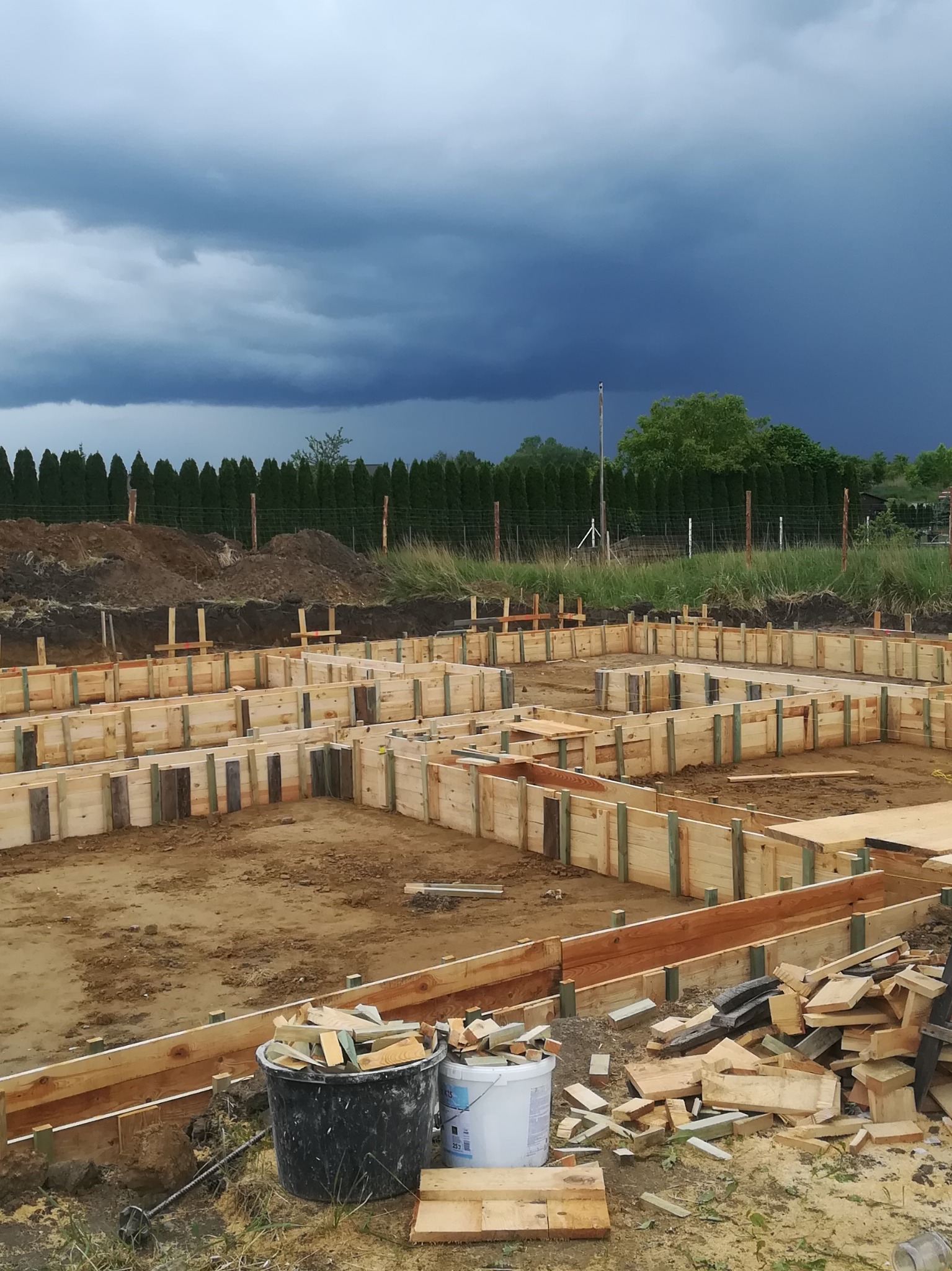 Drewniany szalunek fundamentów domu jednorodzinnego, przygotowany do zalania betonem. W tle ciemne chmury burzowe i rząd tuj.