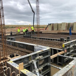 RT-Kraft Sp. z o.o. - Zbrojenie fundamentów z użyciem metalowych szalunków i siatki. W tle pracownicy w żółtych kamizelkach i sterta materiałów budowlanych. Widok na plac budowy w pochmurny dzień.