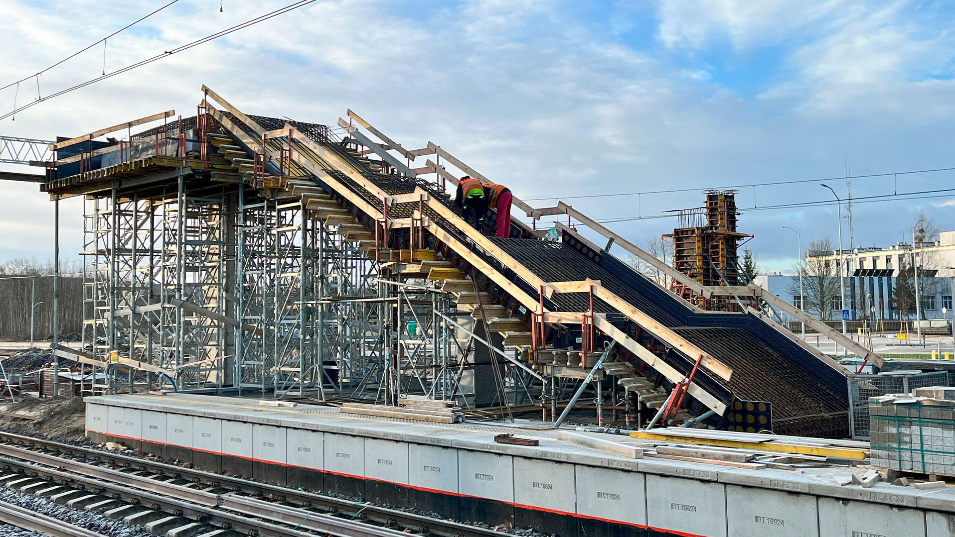 Budowa schodów na peronie kolejowym, widoczne rusztowania, zbrojenia i pracujący robotnicy w odblaskowych kamizelkach. Konstrukcja w trakcie realizacji.