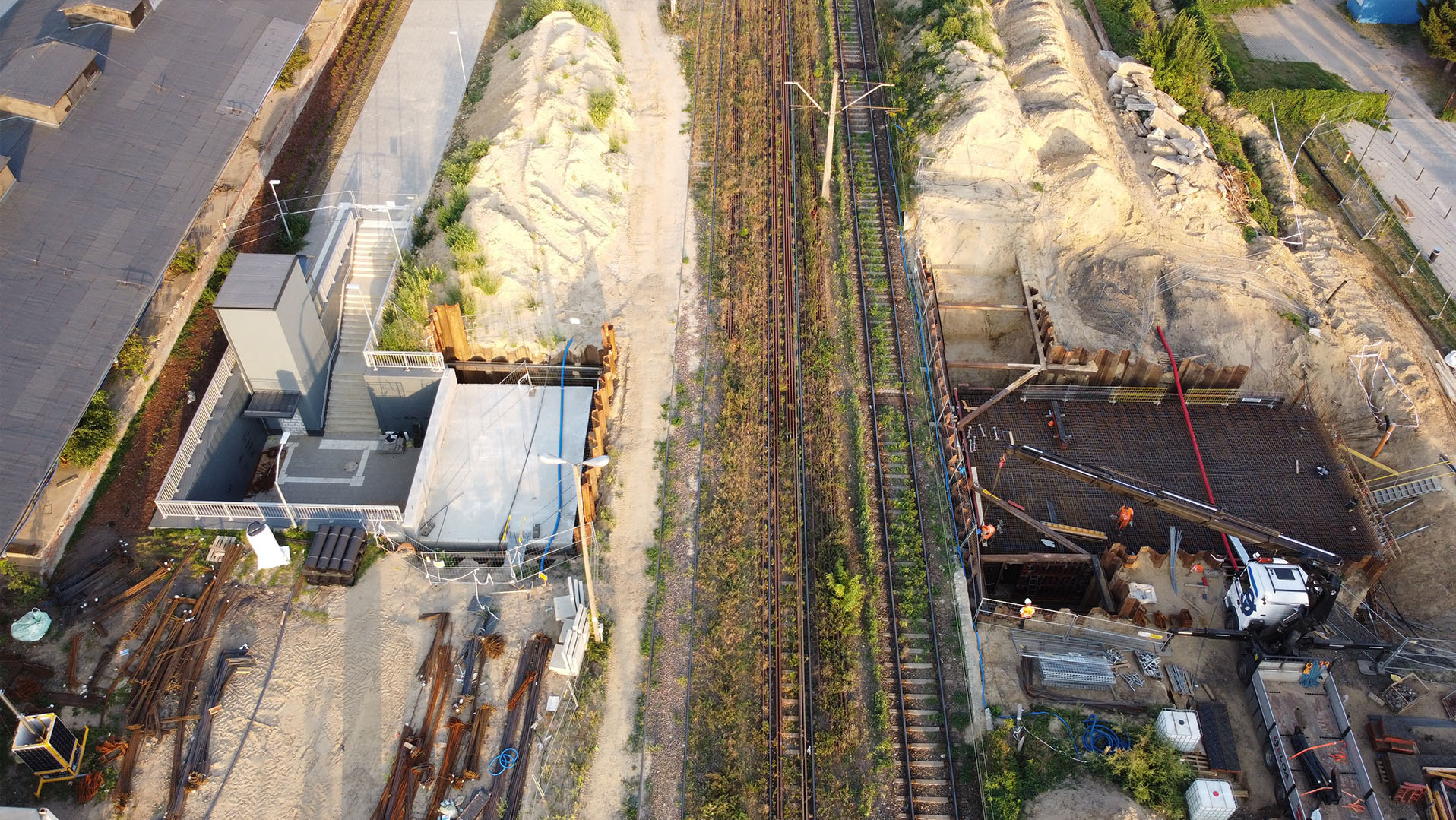 Zdjęcie z drona: budowa fundamentów pod budynek przy torach kolejowych. Widoczne zbrojenia, wykop i ciężarówka. Na drugim planie schody i budynek.