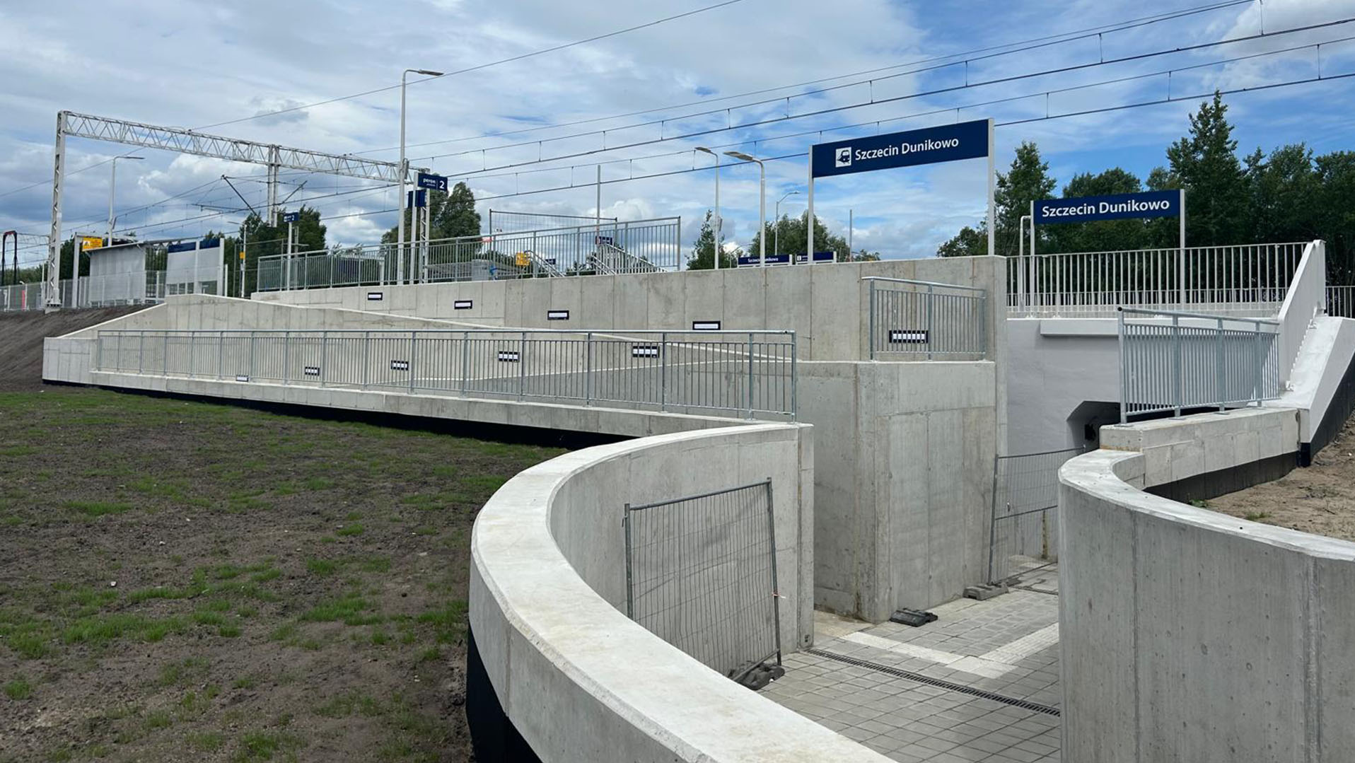 Nowoczesna, betonowa infrastruktura stacji Szczecin Dunikowo z rampami i schodami, otoczona zielenią i błękitnym niebem. Widoczne znaki informacyjne stacji.