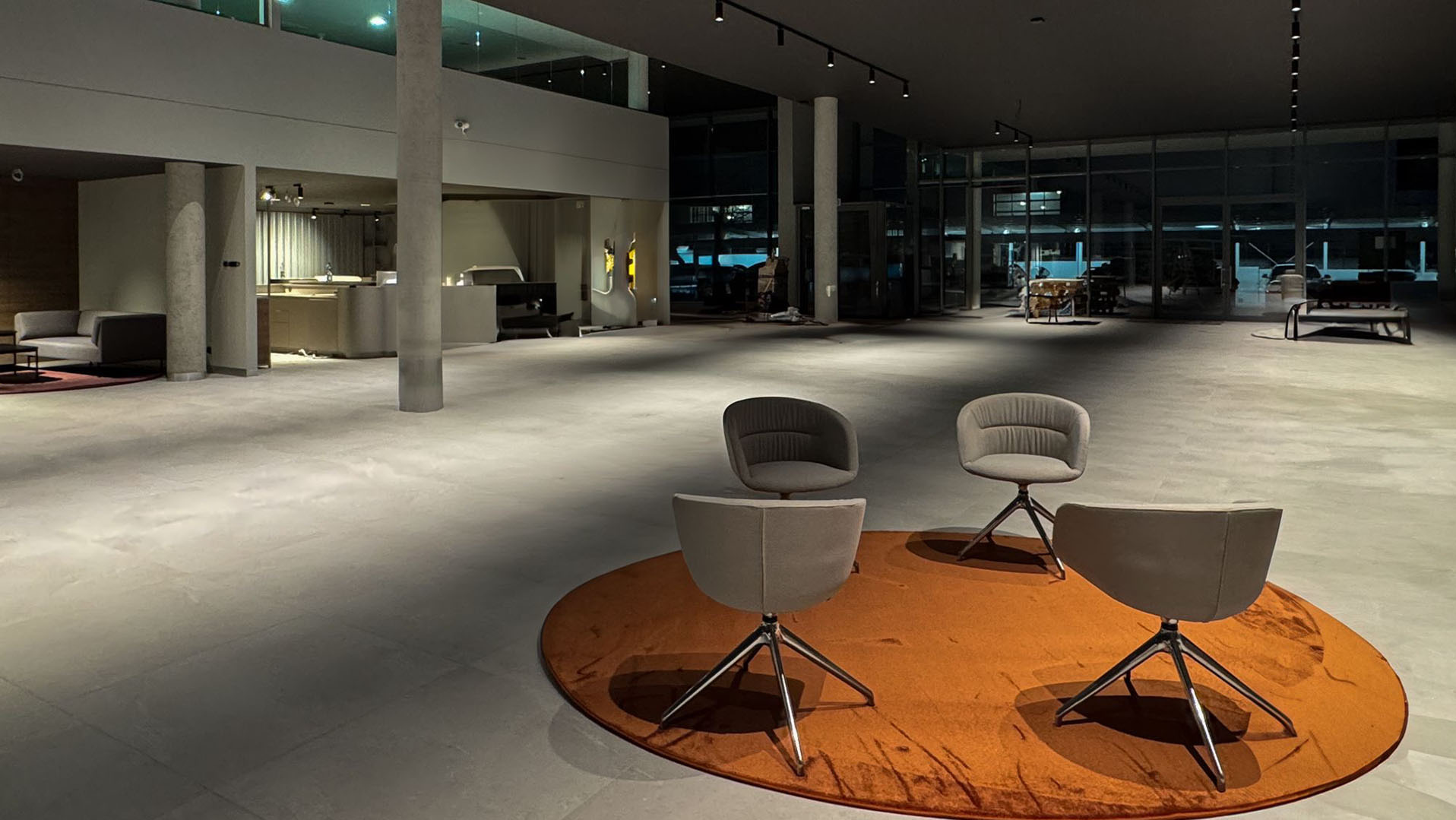 Przestronne wnętrze biurowe z betonową posadzką i kolumnami. Na pierwszym planie okrągły dywan z czterema szarymi krzesłami. W tle widoczne recepcja i przeszklona ściana.