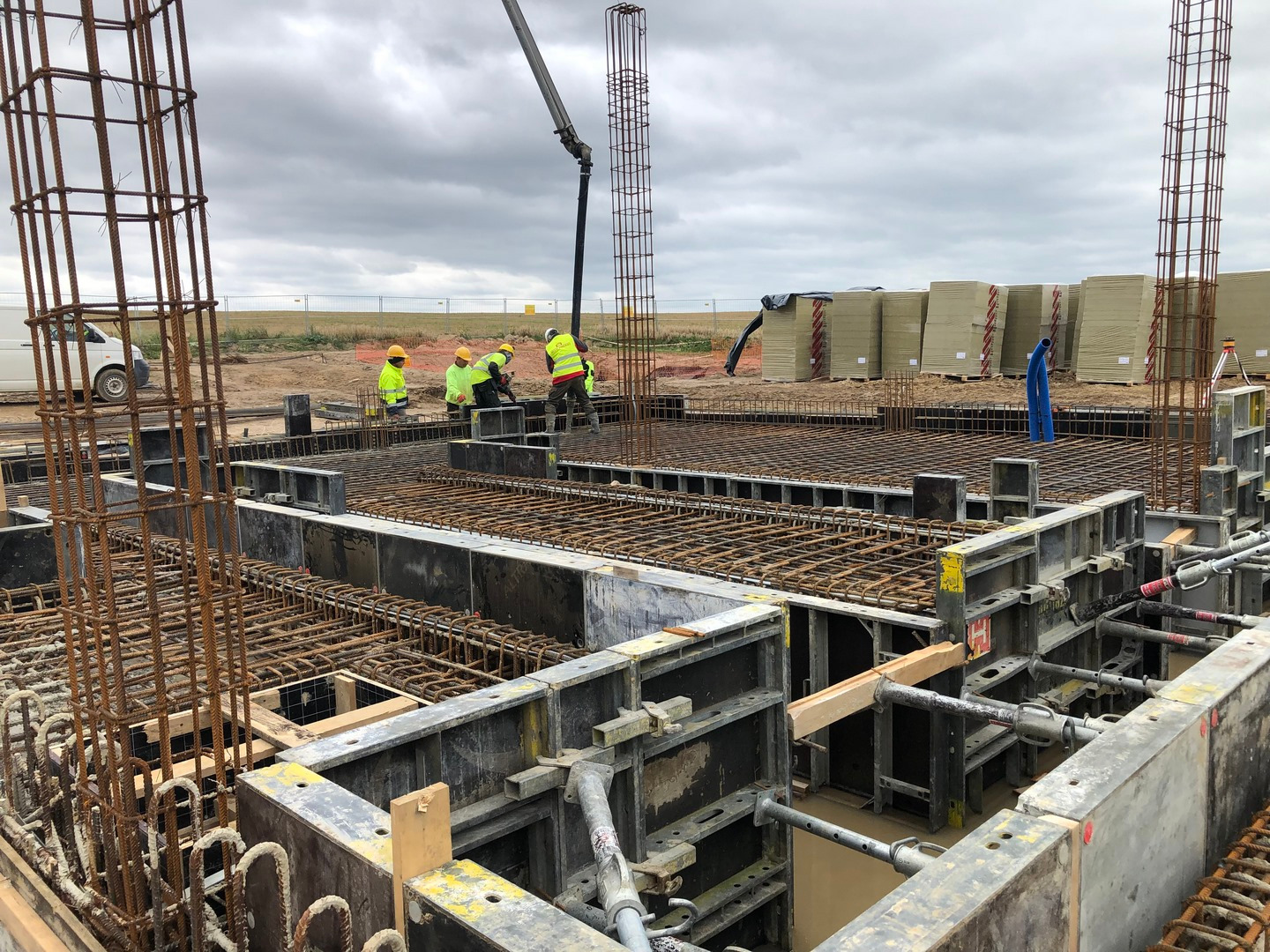 Zbrojenie fundamentów z użyciem metalowych szalunków i siatki. W tle pracownicy w żółtych kamizelkach i sterta materiałów budowlanych. Widok na plac budowy w pochmurny dzień.
