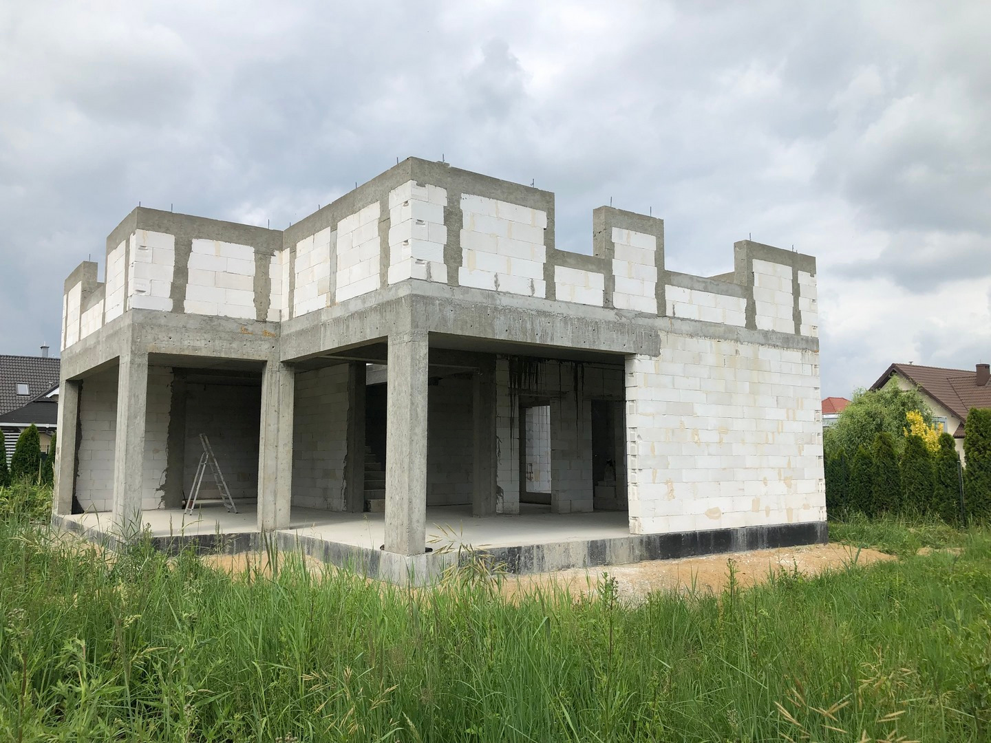 Surowy stan domu z betonu komórkowego w trakcie budowy, widok z zewnątrz na tle zachmurzonego nieba i zielonej trawy. Widoczna drabina wewnątrz konstrukcji.