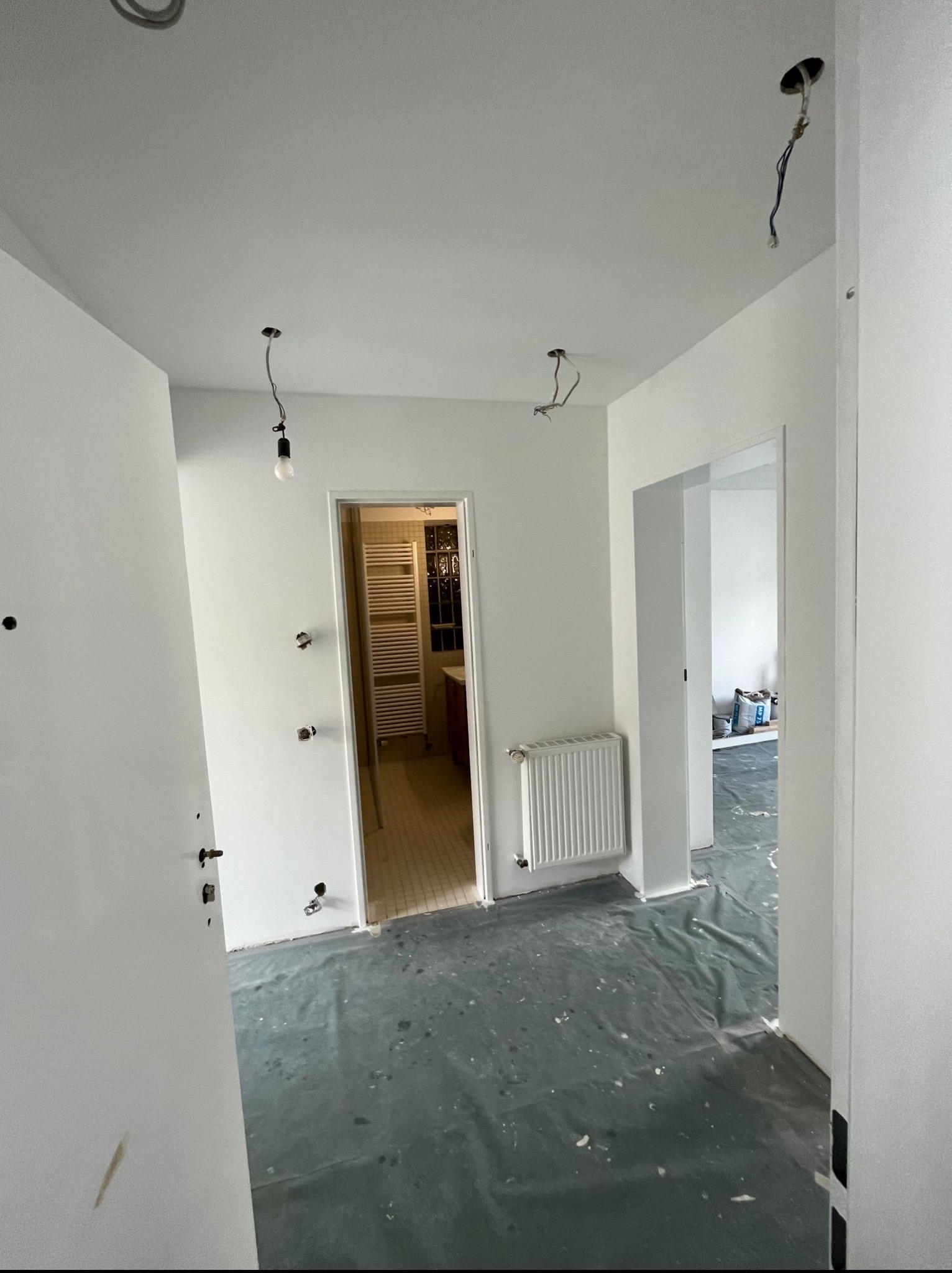 Wnętrze po świeżym malowaniu: białe ściany, zabezpieczona podłoga folią, widoczne instalacje elektryczne i grzejniki. W tle wykończona łazienka z grzejnikiem.