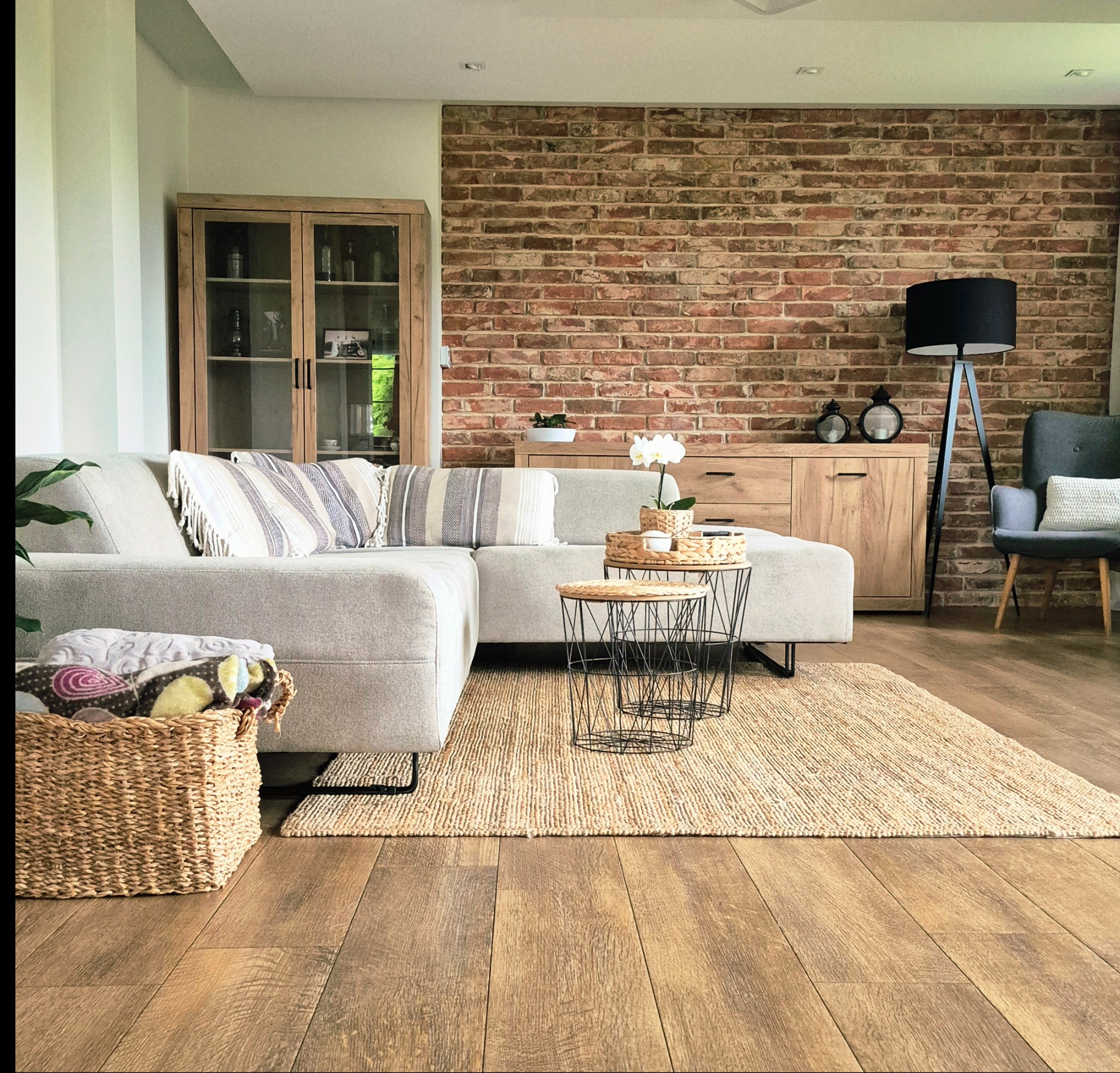 Salon w stylu rustykalnym: ceglana ściana, szara sofa z poduszkami, drewniane meble, dywan z juty i stoliki kawowe z metalowymi nogami.