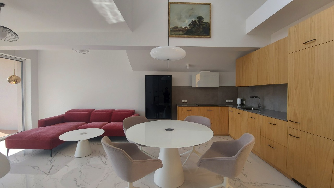 Jasne, nowoczesne wnętrze: czerwona sofa, okrągły stół z krzesłami i drewniana zabudowa kuchenna z szarym blatem. Obraz na ścianie dodaje charakteru.