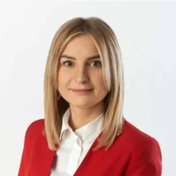 Portret kobiety z blond włosami w czerwonej marynarce na białym tle. Uśmiechnięta, patrzy wprost. Profesjonalny wizerunek.