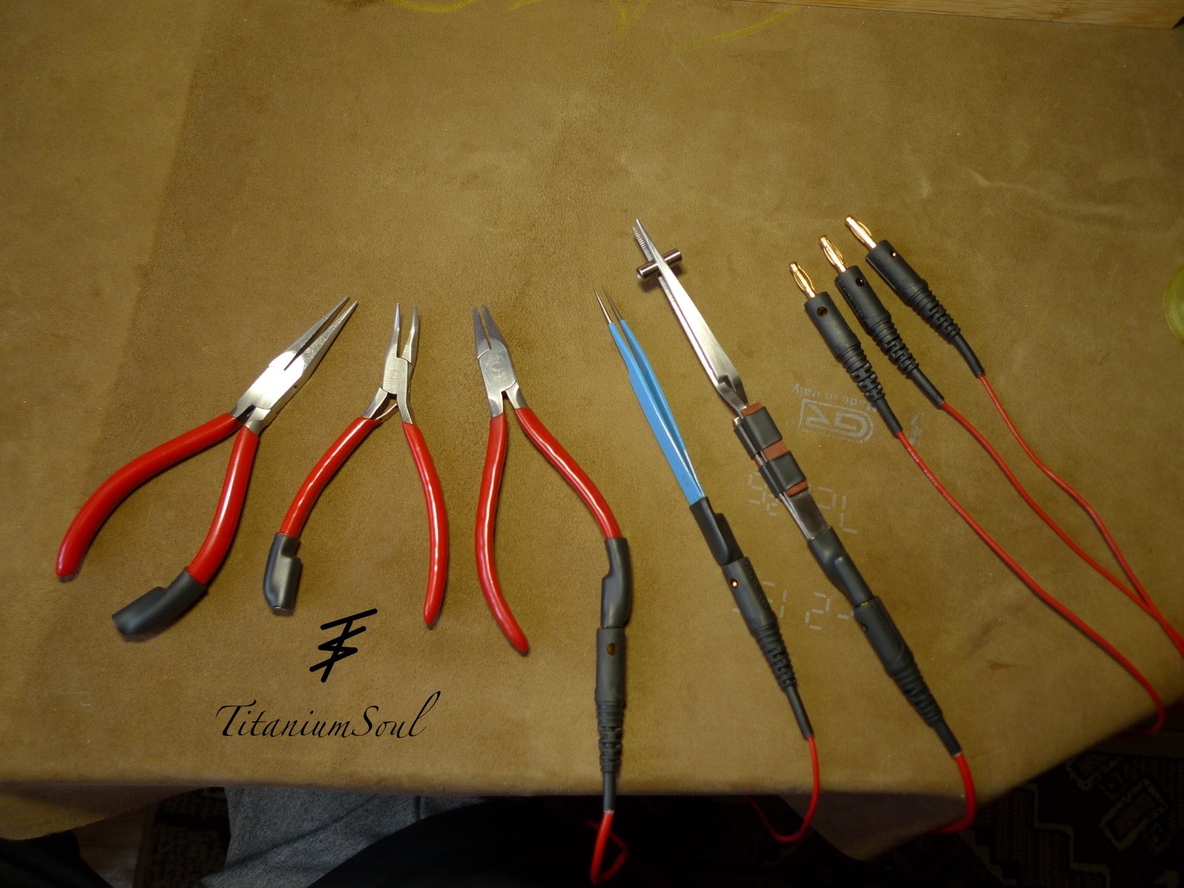 Zestaw narzędzi do precyzyjnych prac spawalniczych: trzy rodzaje szczypiec z czerwonymi uchwytami, niebieska pęseta i przewody z wtykami bananowymi na brązowym tle.