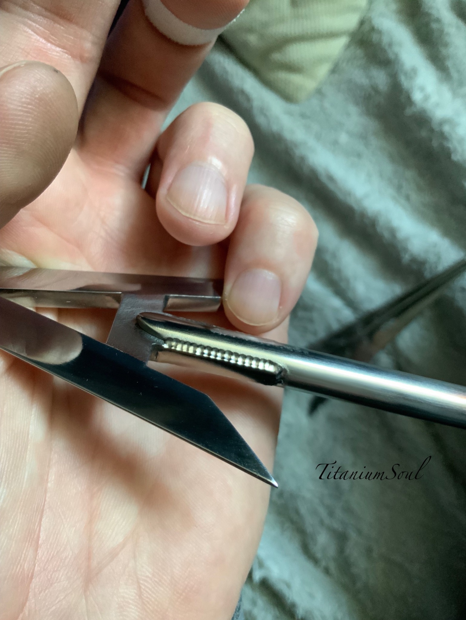 Dłoń trzymająca szczypce spawalnicze z drobnymi ząbkami, zbliżenie na mechanizm, szary materiał w tle. Narzędzie precyzyjne dla spawacza.