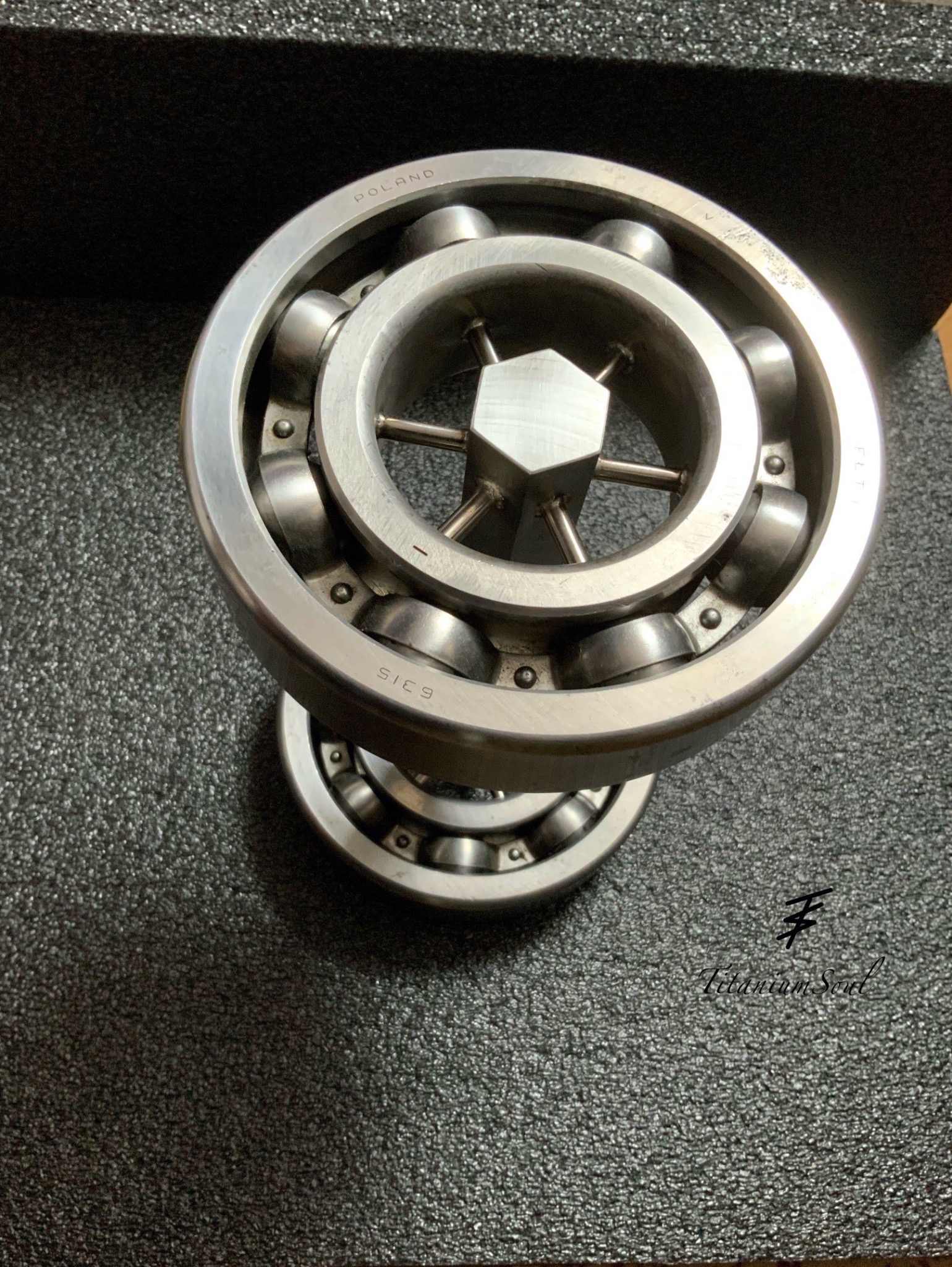 Dwa łożyska kulkowe FLT 6315 z napisem POLAND, ułożone jedno na drugim, na szarym tle. Widoczne detale konstrukcji i oznaczenia.