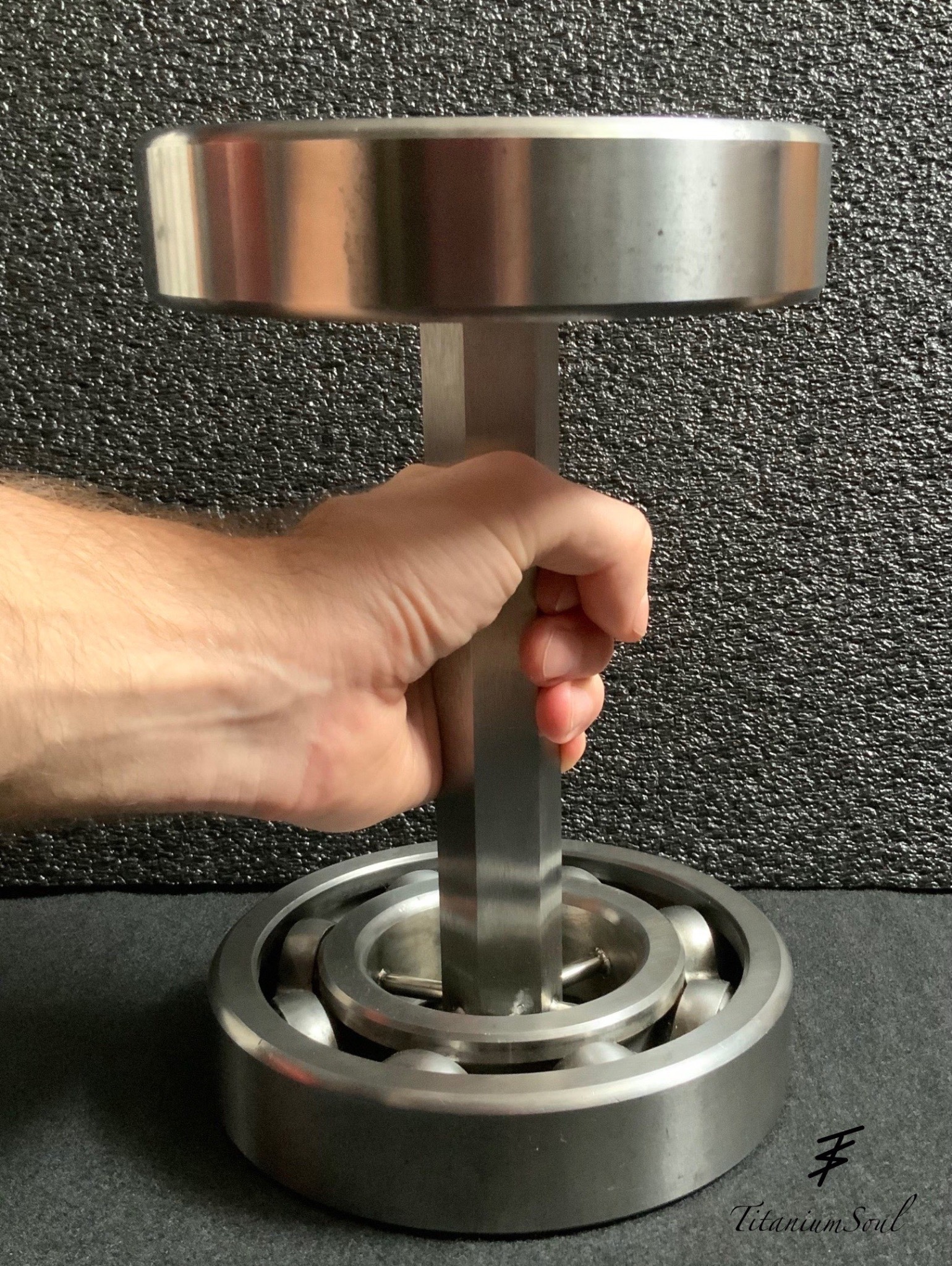 Metalowa konstrukcja spawana, przypominająca hantlę, trzymana w dłoni na tle szarej ściany. Widoczne spawy i elementy metalowe.