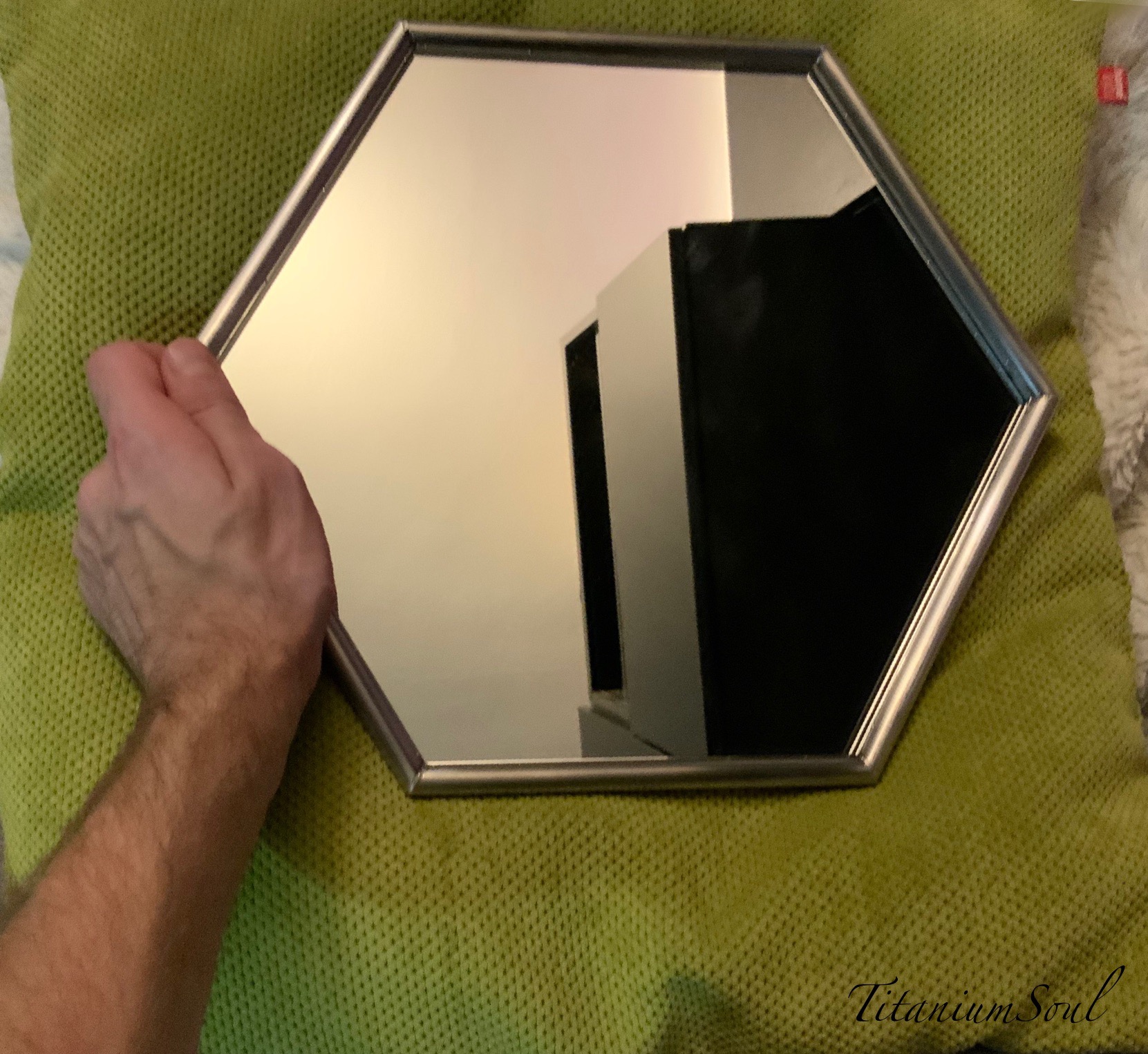 Srebrna, heksagonalna rama lustra w dłoni, odbijająca wnętrze. Leży na zielonej poduszce, akcentując precyzję wykonania ramy.