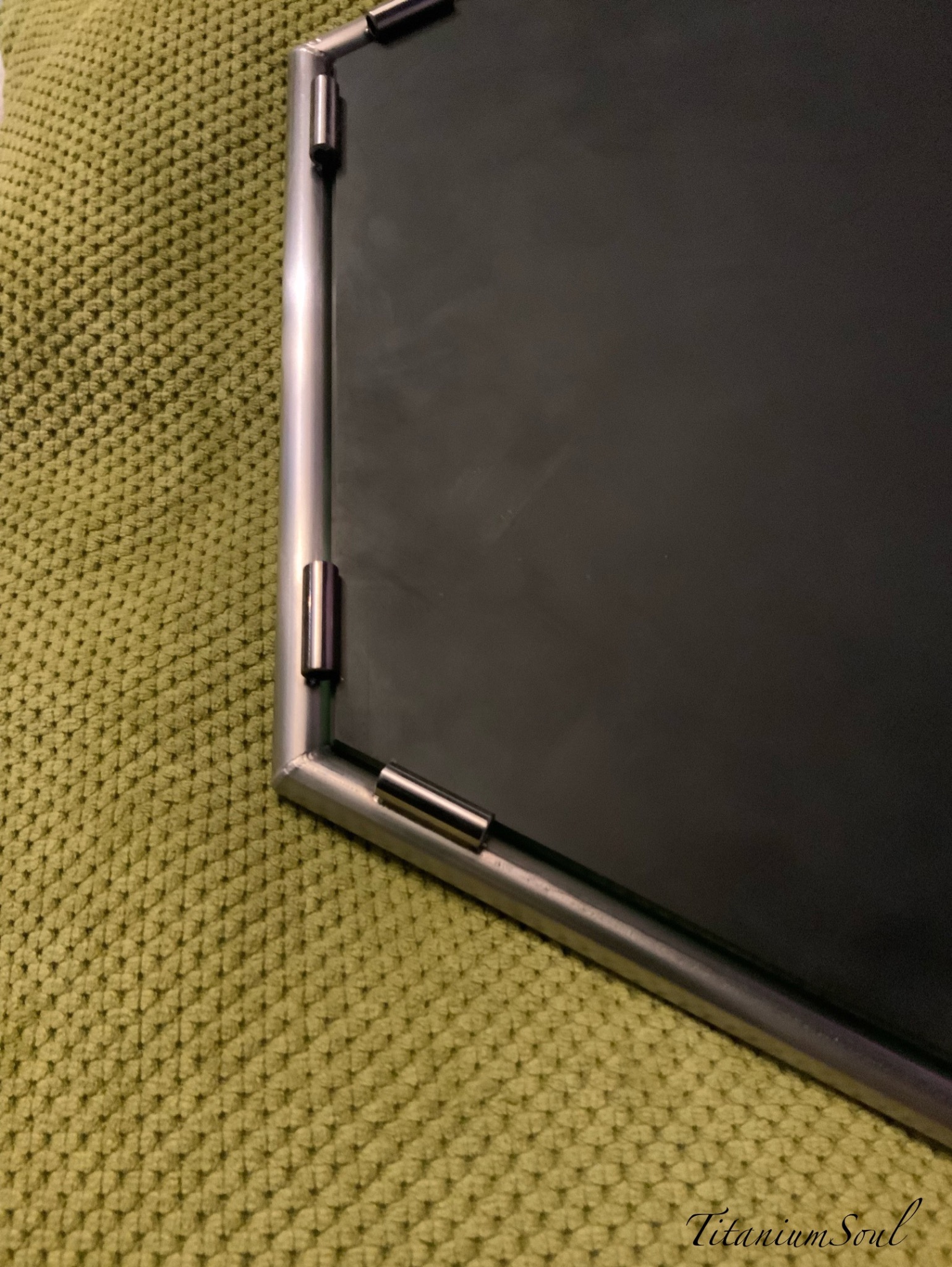 Fragment metalowej ramy z łącznikami, leżącej na zielonej, teksturowanej powierzchni. Widoczny spaw na rogu ramy. Precyzyjne wykonanie.