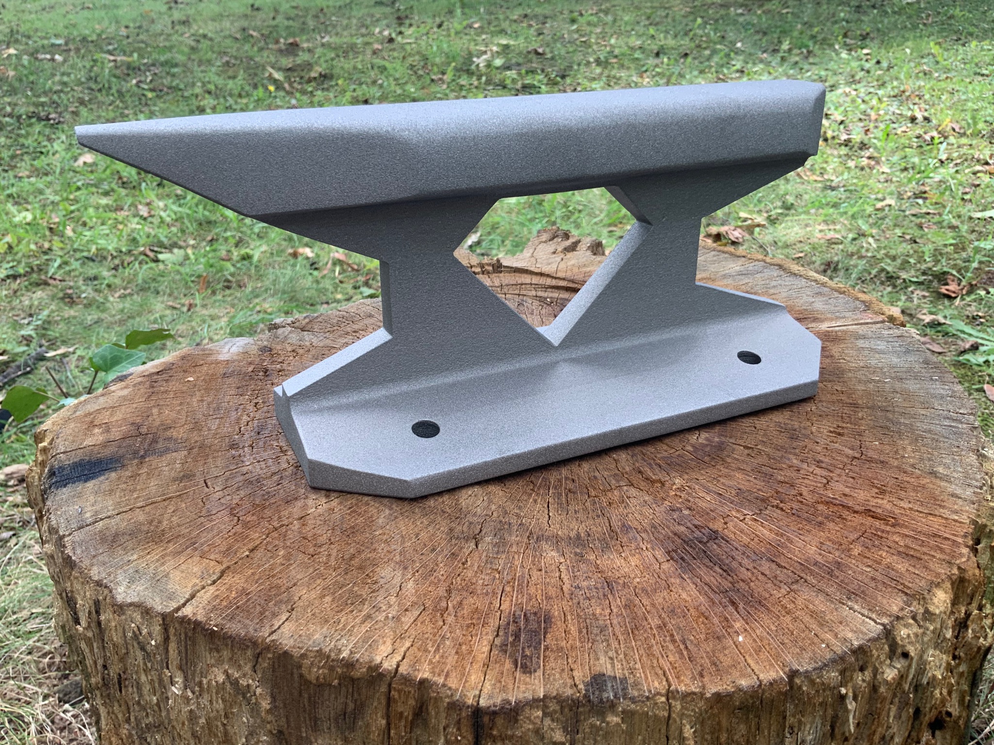 Szara, spawana kowadło na pniu drzewa w plenerze. Widoczne ślady obróbki metalu i otwory montażowe. Kowadło o nowoczesnym designie.