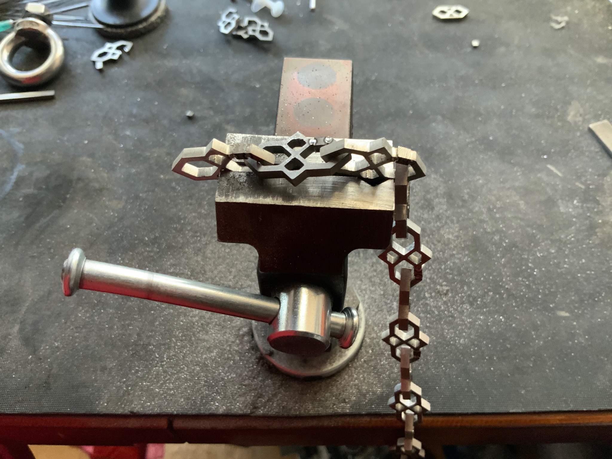 Metalowy łańcuch w imadle, detale spawania ogniw. Widoczne narzędzia i drobne elementy metalowe na warsztacie. Obróbka metalu w trakcie realizacji.