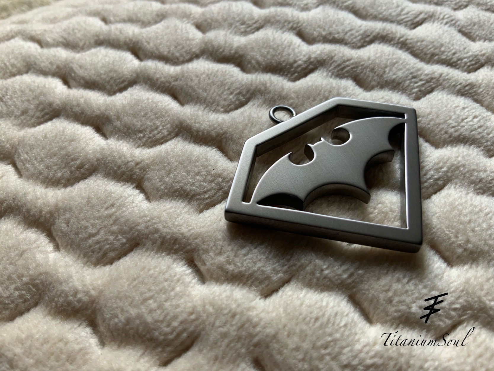 Metalowy brelok w kształcie nietoperza w ramce, leżący na miękkiej, kremowej tkaninie. Widoczny sygnatura firmy 'Titanium Soul'.