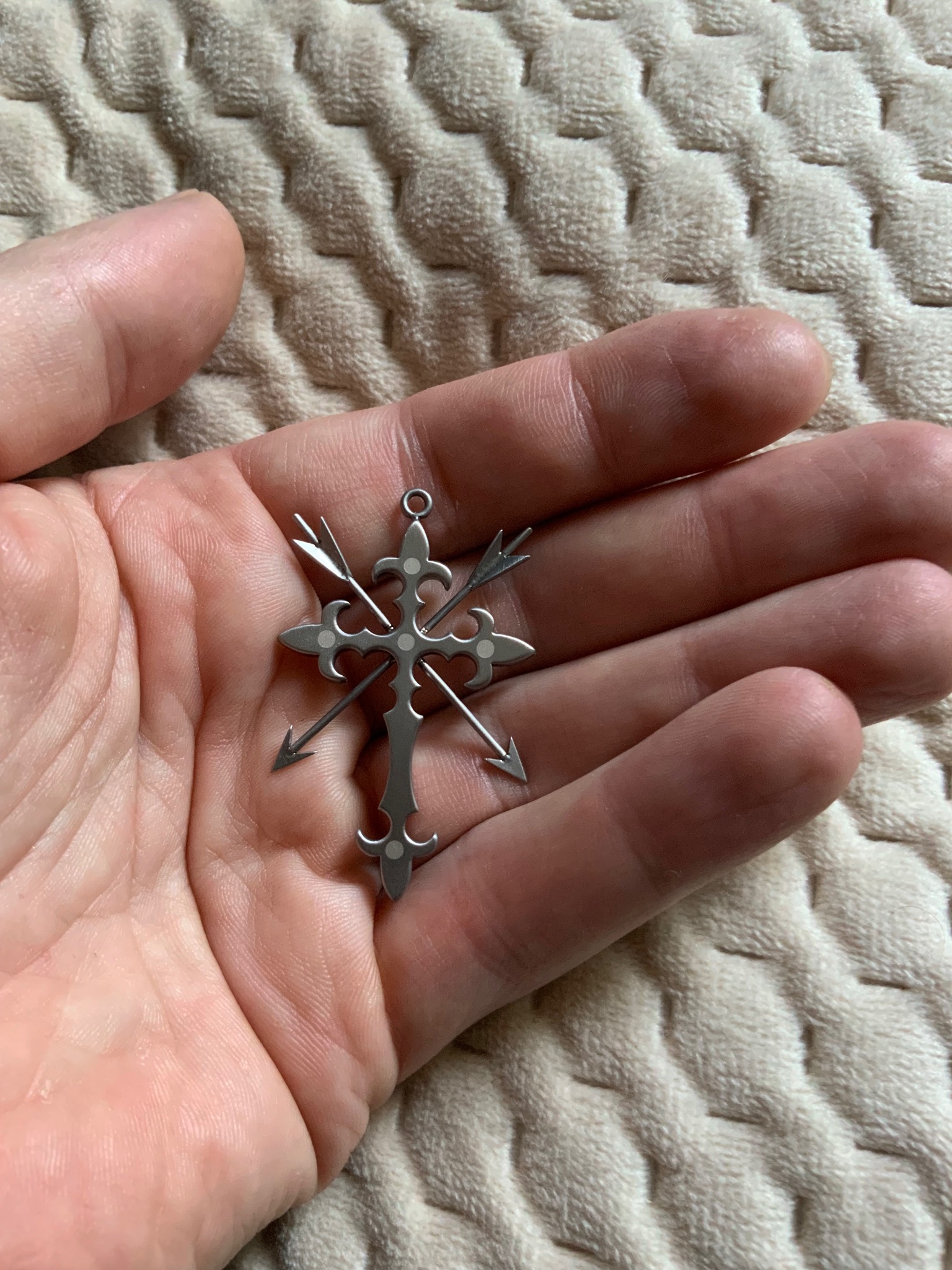 Metalowy krzyżyk z motywem strzał w dłoni na tle teksturowanej tkaniny. Krzyżyk ma ozdobne detale i srebrny kolor.