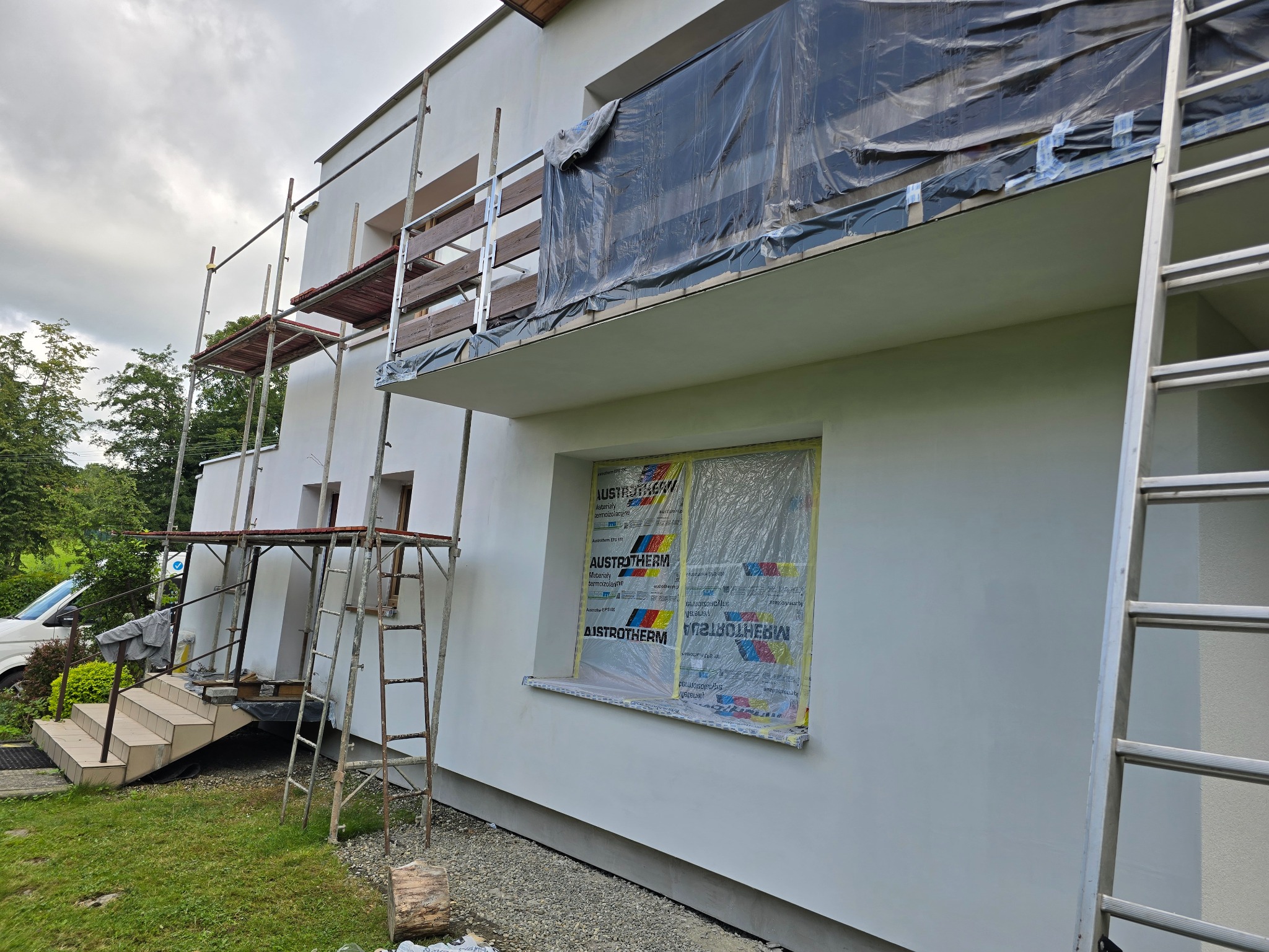 Elewacja domu w trakcie prac: świeżo otynkowana ściana, okno zabezpieczone folią Austrotherm, rusztowania i drabina. Widoczne fragmenty zieleni i nieba.