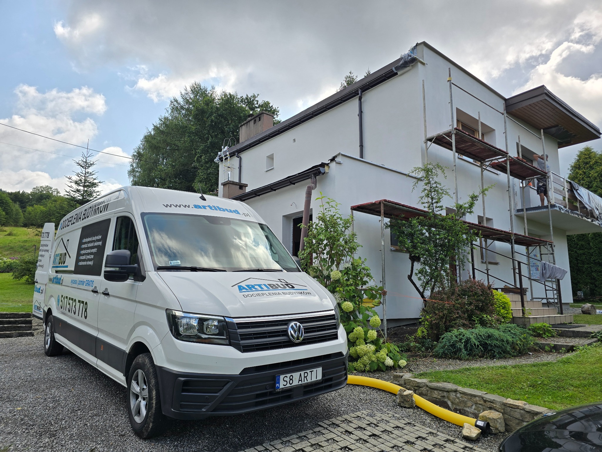 Biała elewacja domu po renowacji w Kiczycach, z widocznym rusztowaniem i samochodem firmy Artibud. Żółty wąż do ocieplania leży na kostce brukowej.