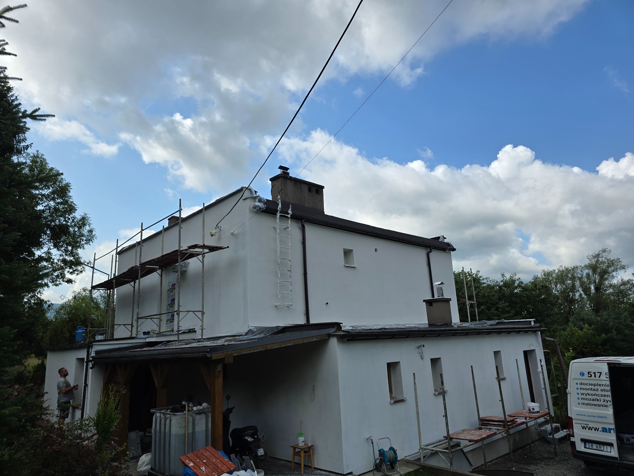 Ocieplony dom z rusztowaniem i drabiną. Częściowo widoczny pracownik malujący elewację. Biała elewacja na tle błękitnego nieba z chmurami.