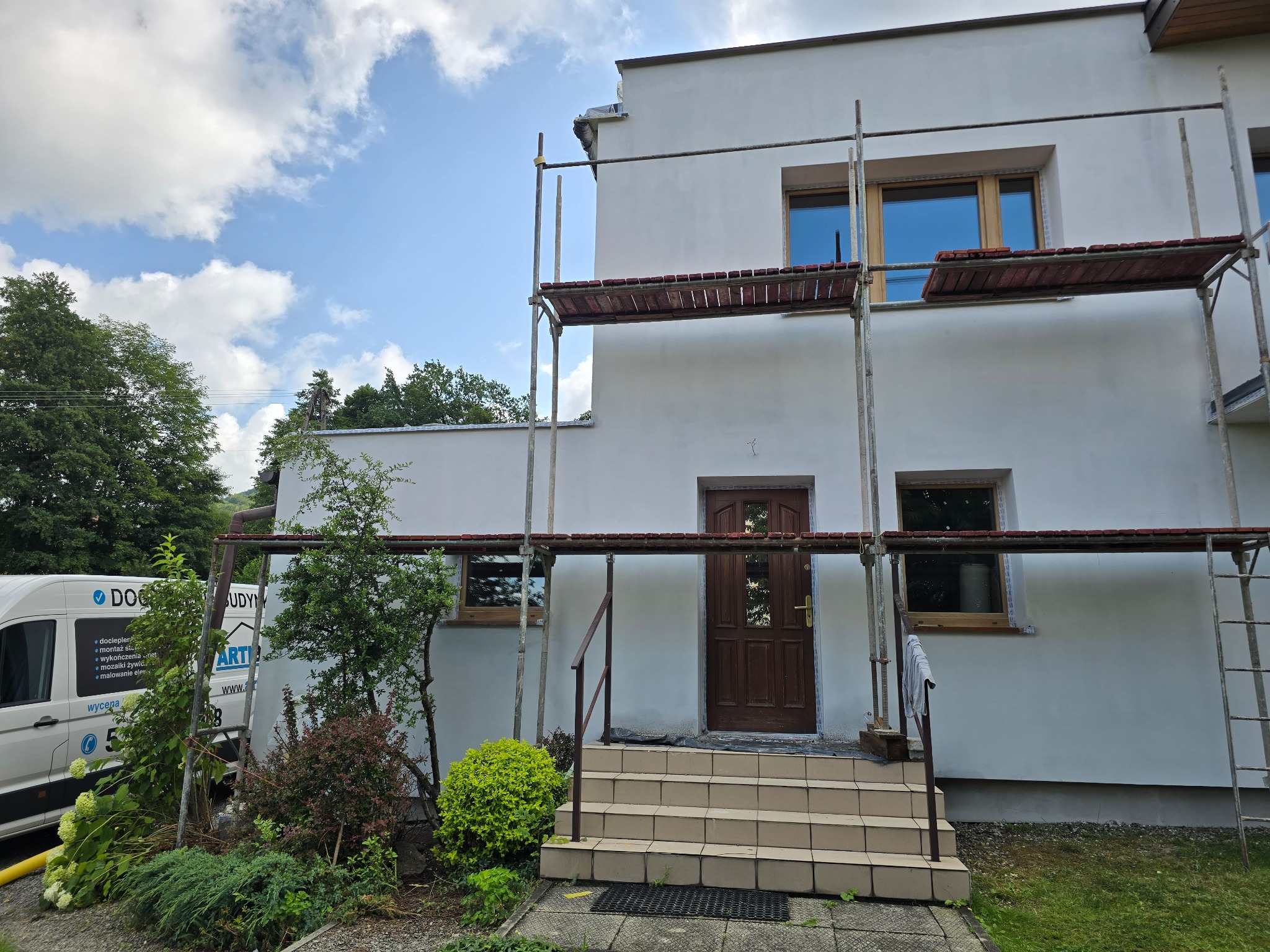 Biały dom jednorodzinny w trakcie prac elewacyjnych, widoczne rusztowania i świeżo pomalowana ściana. Częściowo zasłonięty firmowym vanem z logo.