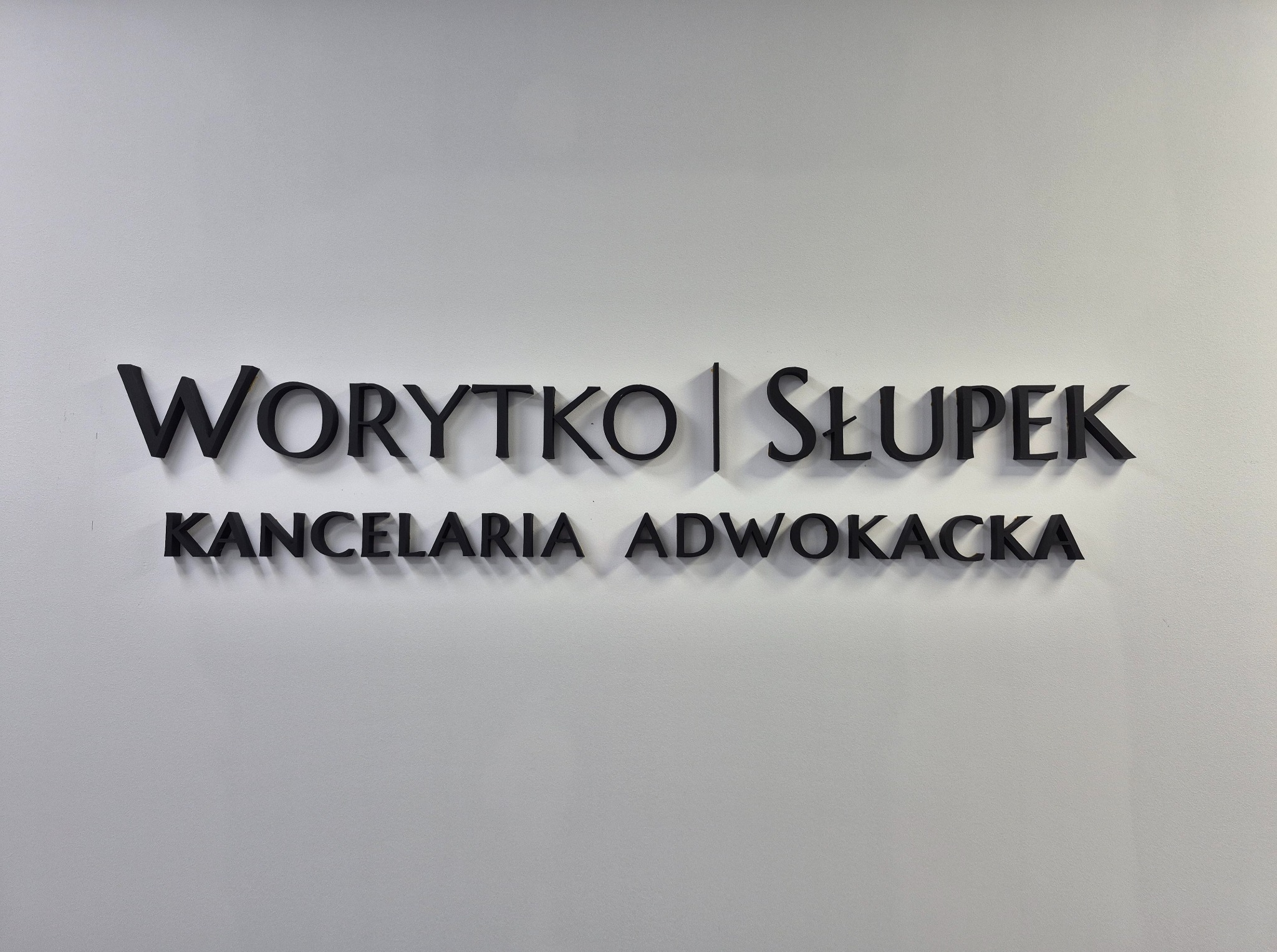 Tablica informacyjna: czarne litery na białej ścianie z nazwą 'Worytko | Słupek Kancelaria Adwokacka' w minimalistycznym stylu.