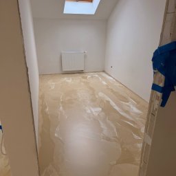 Damian Karasiewicz - Jasna, błyszcząca podłoga w trakcie wylewania, widoczna przez otwarte drzwi. W tle biały grzejnik i okno dachowe. Niebieska taśma zabezpieczająca na framudze.