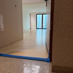 Damian Karasiewicz - Jasna, lśniąca podłoga w trakcie prac wykończeniowych, zabezpieczona niebieską taśmą malarską przy ścianach i futrynach. Widok z niskiej perspektywy na wnętrze mieszkania.