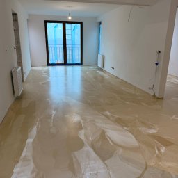 Damian Karasiewicz - Beżowa, błyszcząca podłoga w przestronnym, jasnym pokoju z dużym oknem balkonowym zabezpieczonym folią. Widoczne niedokończone prace wykończeniowe.