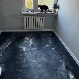 Damian Karasiewicz - Czarno-biała, błyszcząca podłoga z żywicy w pokoju z grzejnikiem i oknem, na którym siedzi czarny kot. Widok na budynek mieszkalny za oknem.