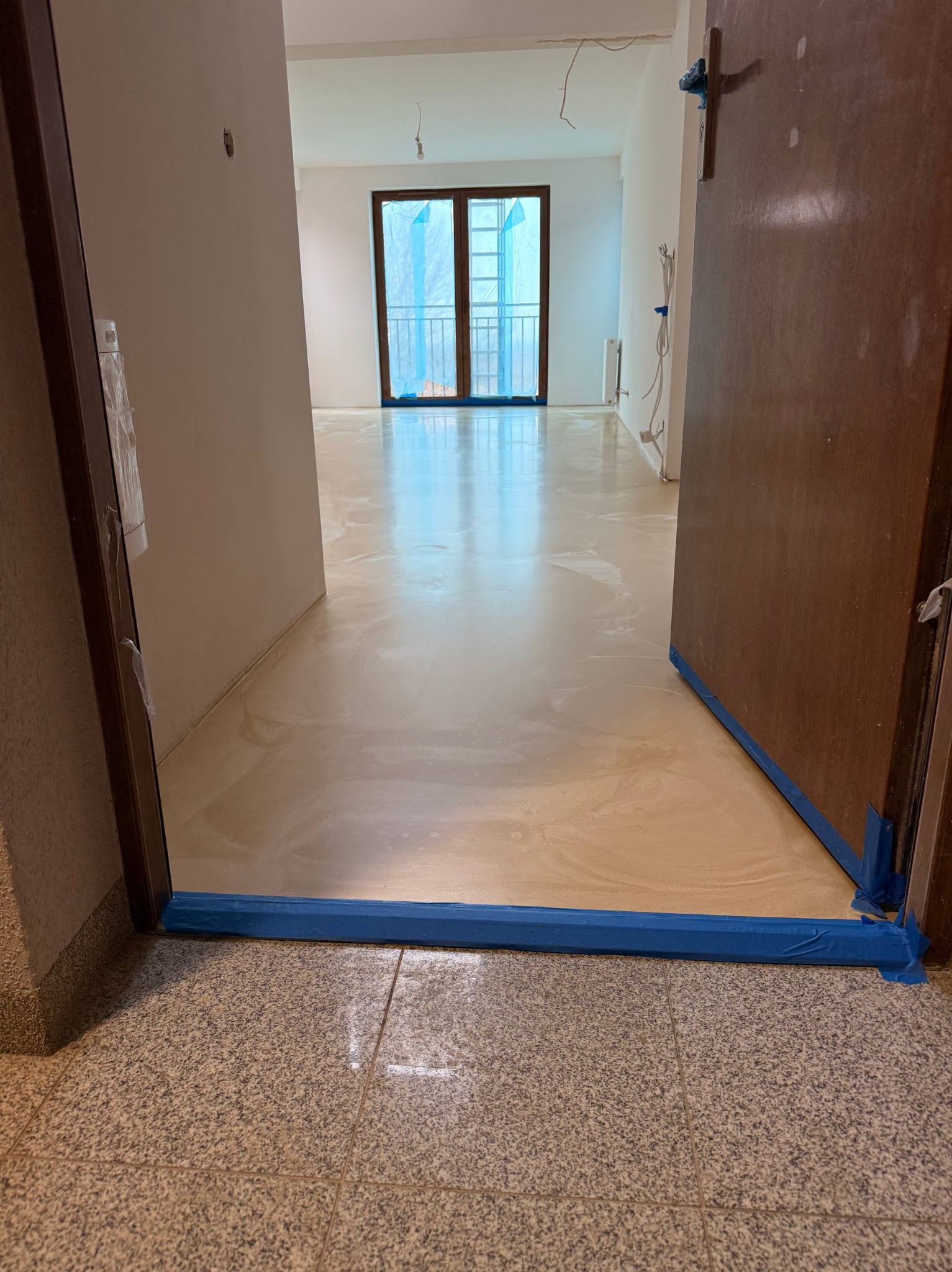 Jasna, błyszcząca posadzka w wykończonym wnętrzu, zabezpieczona niebieską taśmą malarską przy progu i drzwiach. Widok z perspektywy wejścia, okno w tle.