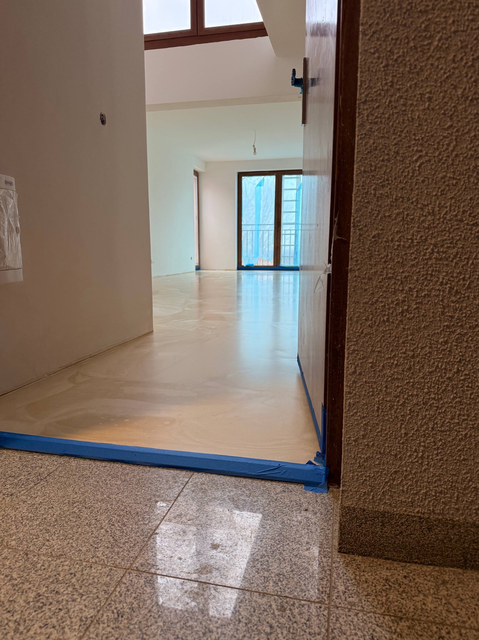 Jasna, lśniąca podłoga w trakcie prac wykończeniowych, zabezpieczona niebieską taśmą malarską przy ścianach i futrynach. Widok z niskiej perspektywy na wnętrze mieszkania.