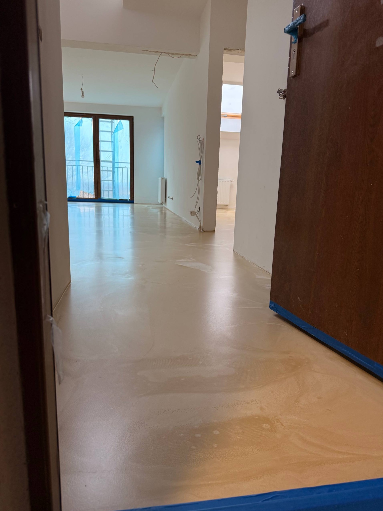 Jasna, beżowa posadzka żywiczna w mieszkaniu po remoncie, zabezpieczona niebieską taśmą malarską, widok z korytarza w Szczecinie. Gładka powierzchnia, naturalne światło.