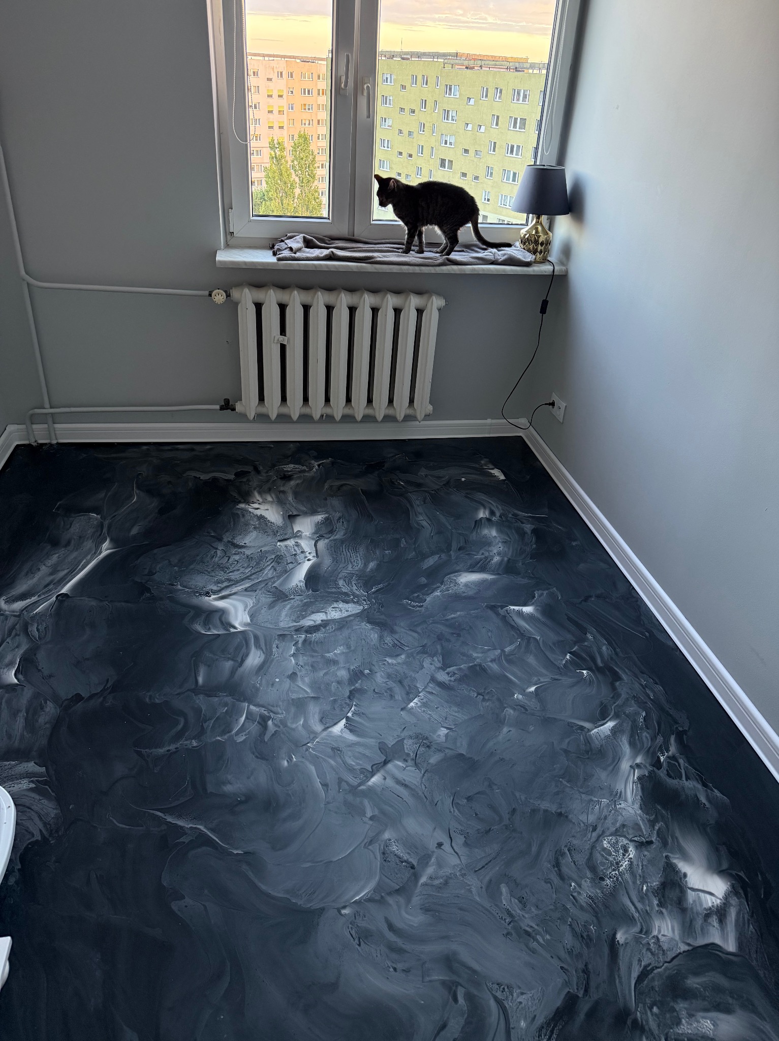 Czarno-biała, błyszcząca podłoga z żywicy w pokoju z grzejnikiem i oknem, na którym siedzi czarny kot. Widok na budynek mieszkalny za oknem.