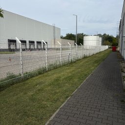 BARTOSZ SZYMAŃSKI - Świeżo skoszona trawa wzdłuż chodnika z kostki brukowej i białego ogrodzenia z drutem kolczastym, na tle szarego magazynu z rampami. Widoczny zbiornik.