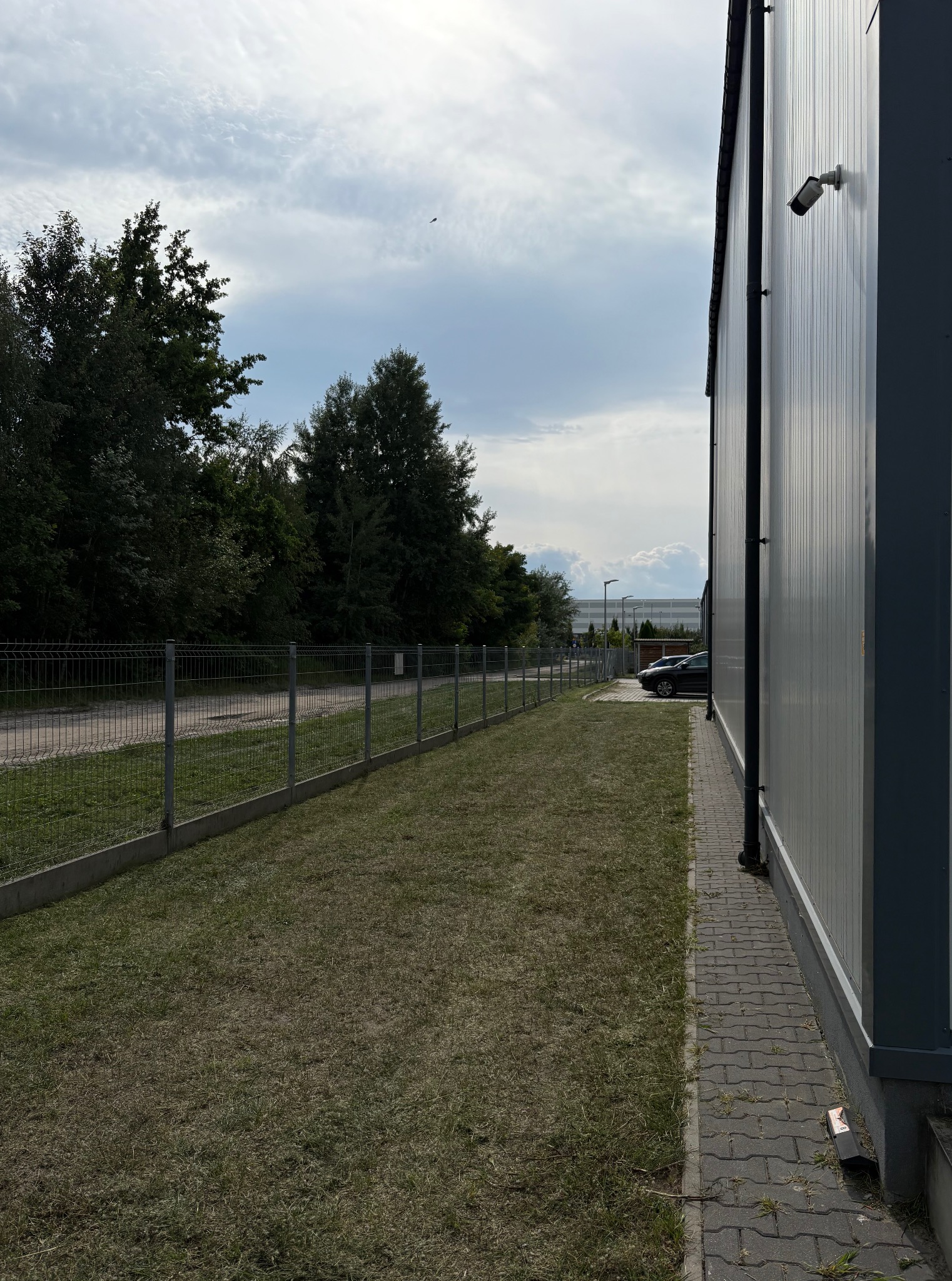 Trawa przy budynku z metalową elewacją i ogrodzeniem panelowym. Widoczny fragment parkingu i drzewa w tle. Nierównomiernie skoszona trawa.