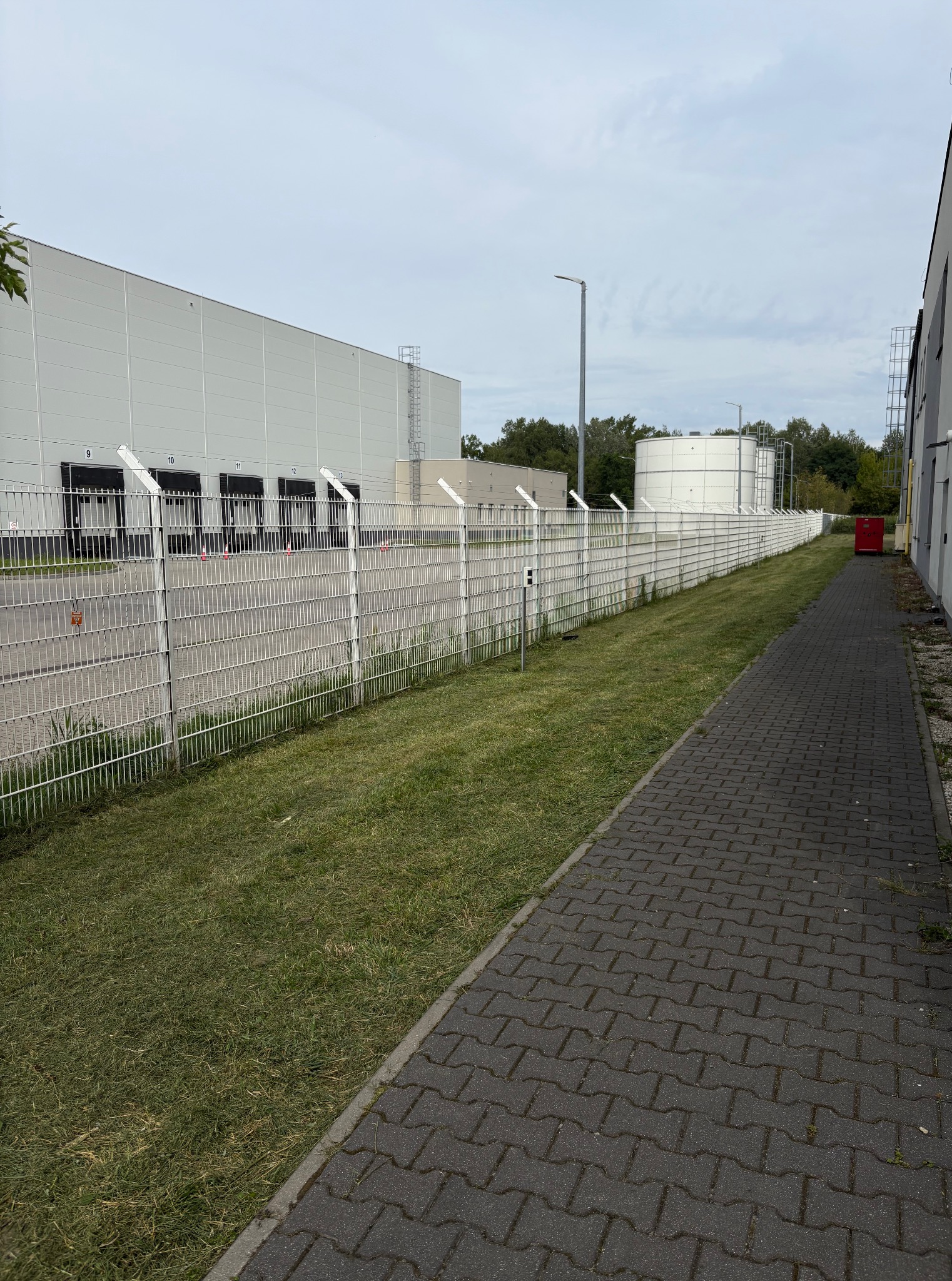 Świeżo skoszona trawa wzdłuż chodnika z kostki brukowej i białego ogrodzenia z drutem kolczastym, na tle szarego magazynu z rampami. Widoczny zbiornik.