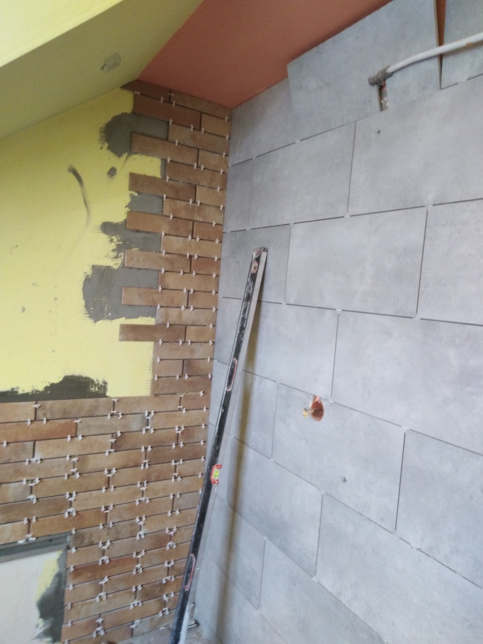 Układanie płytek na ścianie w trakcie remontu. Widoczne płytki w dwóch kolorach i poziomica. Część ściany pomalowana na żółto.