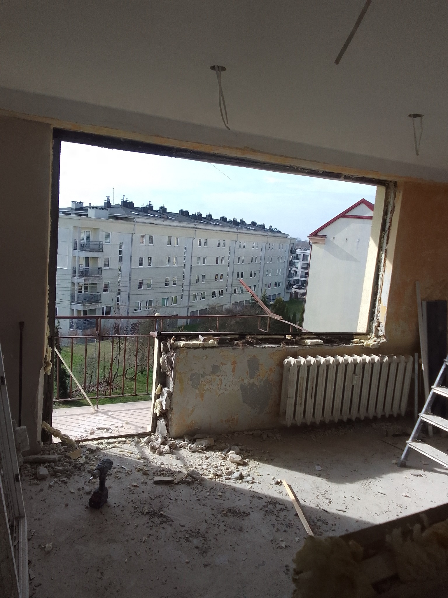 Demontaż okna balkonowego w bloku. Widoczne gruz, stara instalacja grzewcza i narzędzia. W tle budynki mieszkalne.