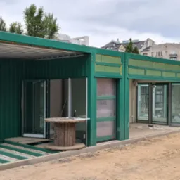 Budowa modułowych pawilonów handlowych z zielonej konstrukcji stalowej, widoczne szklane drzwi i drewniana szpula. Budynki w tle, pochmurne niebo.
